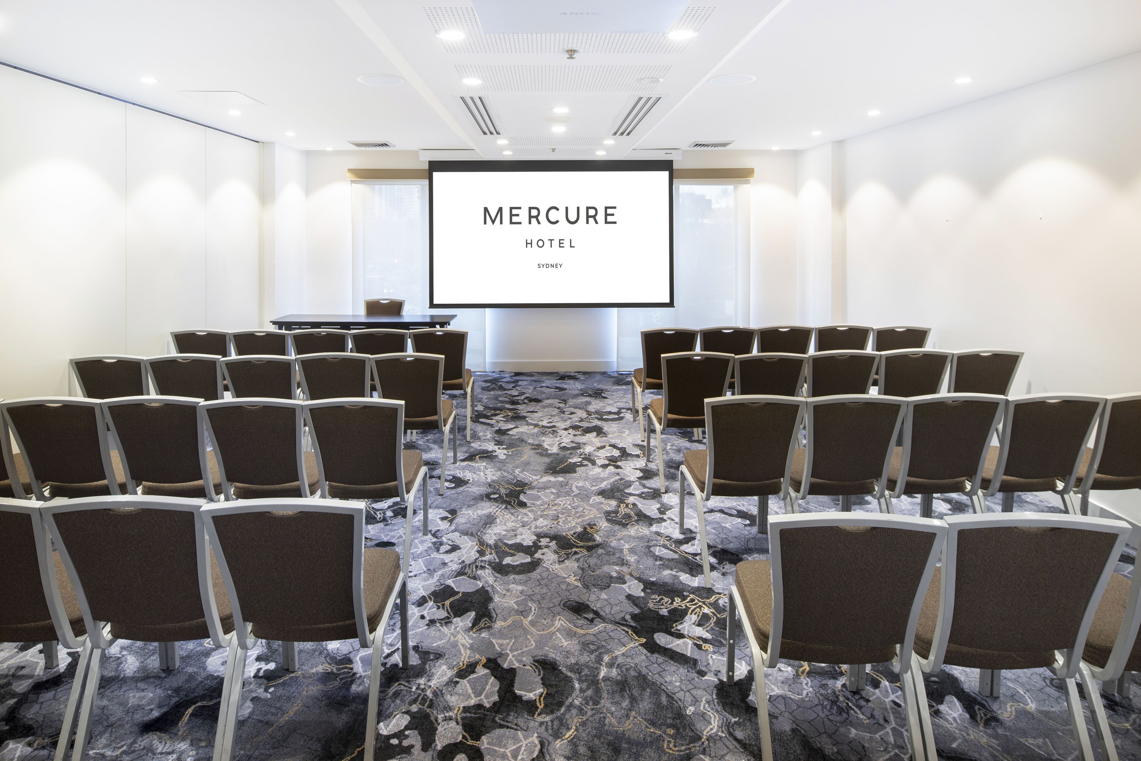 Een vergaderzaal met rijen stoelen, een groot scherm waarop 'Mercure Hotel Sydney' staat en een kamerbreed tapijt met een patroon.