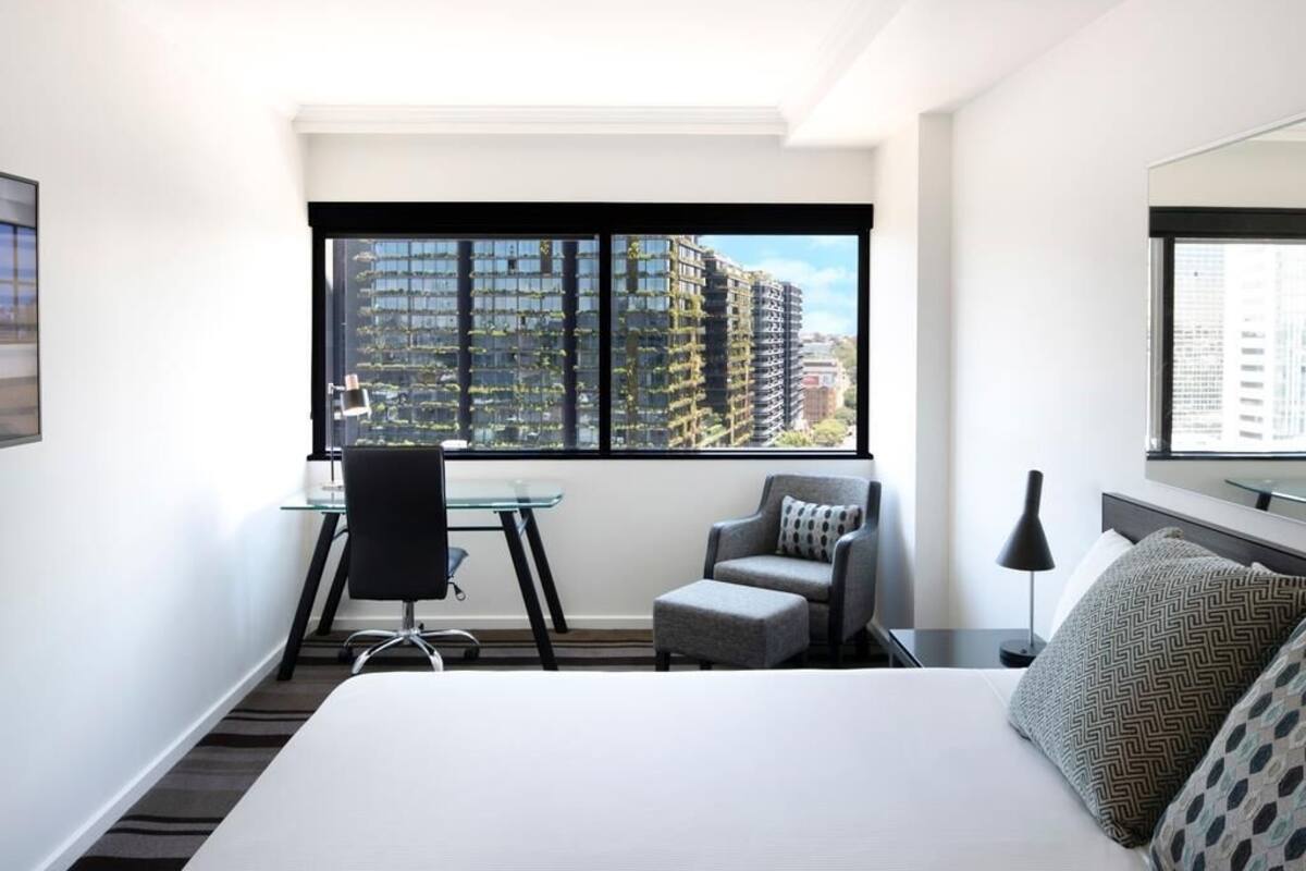 A hotel room with a bed, a desk, a chair, a small table, a lamp, and a large window with a city view.