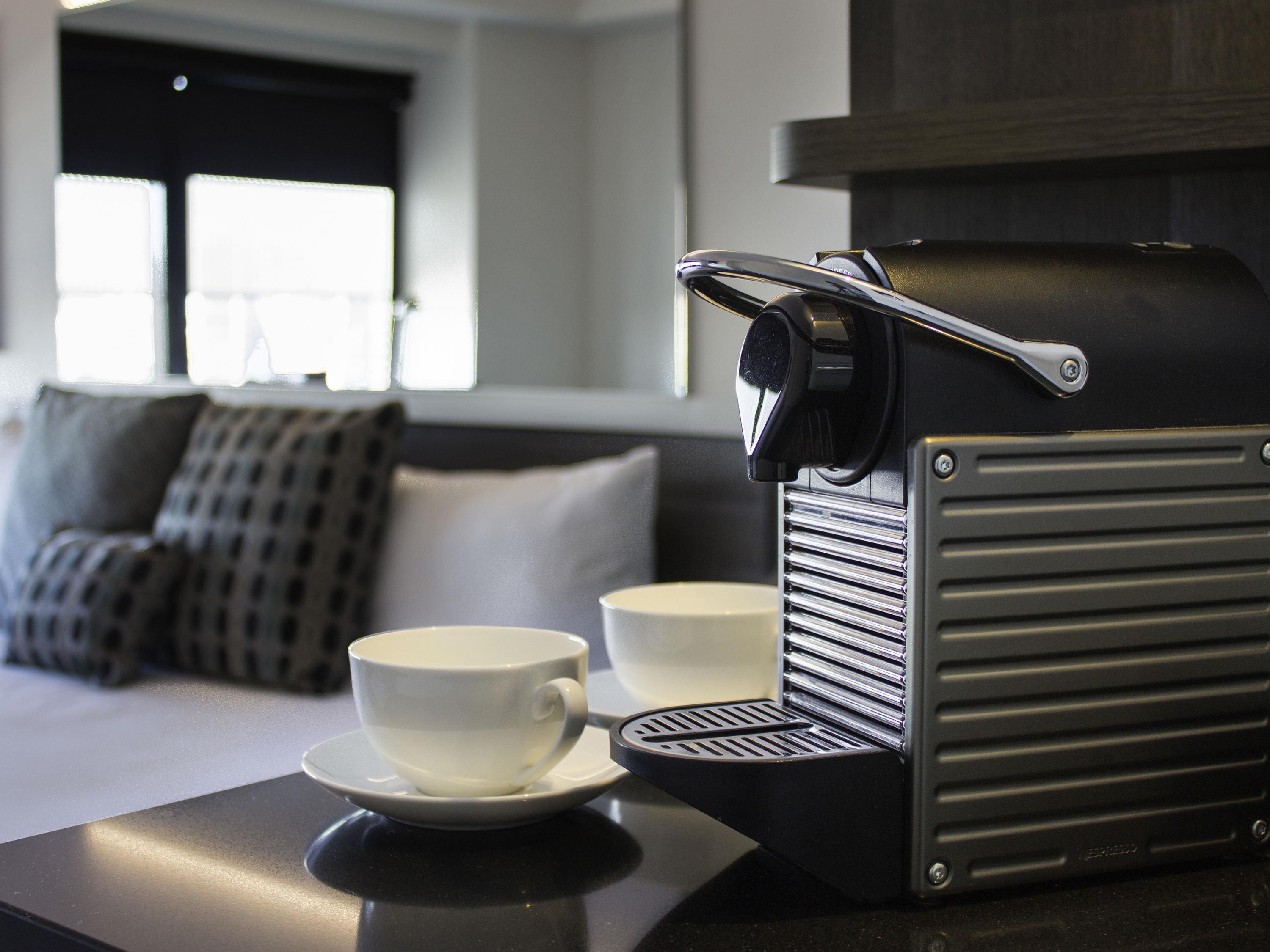 A coffee machine with a cup on a table, a sofa with pillows, and a window with blinds in a room.