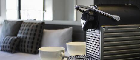 A coffee machine with a cup on a table, a sofa with pillows, and a window with blinds in a room.