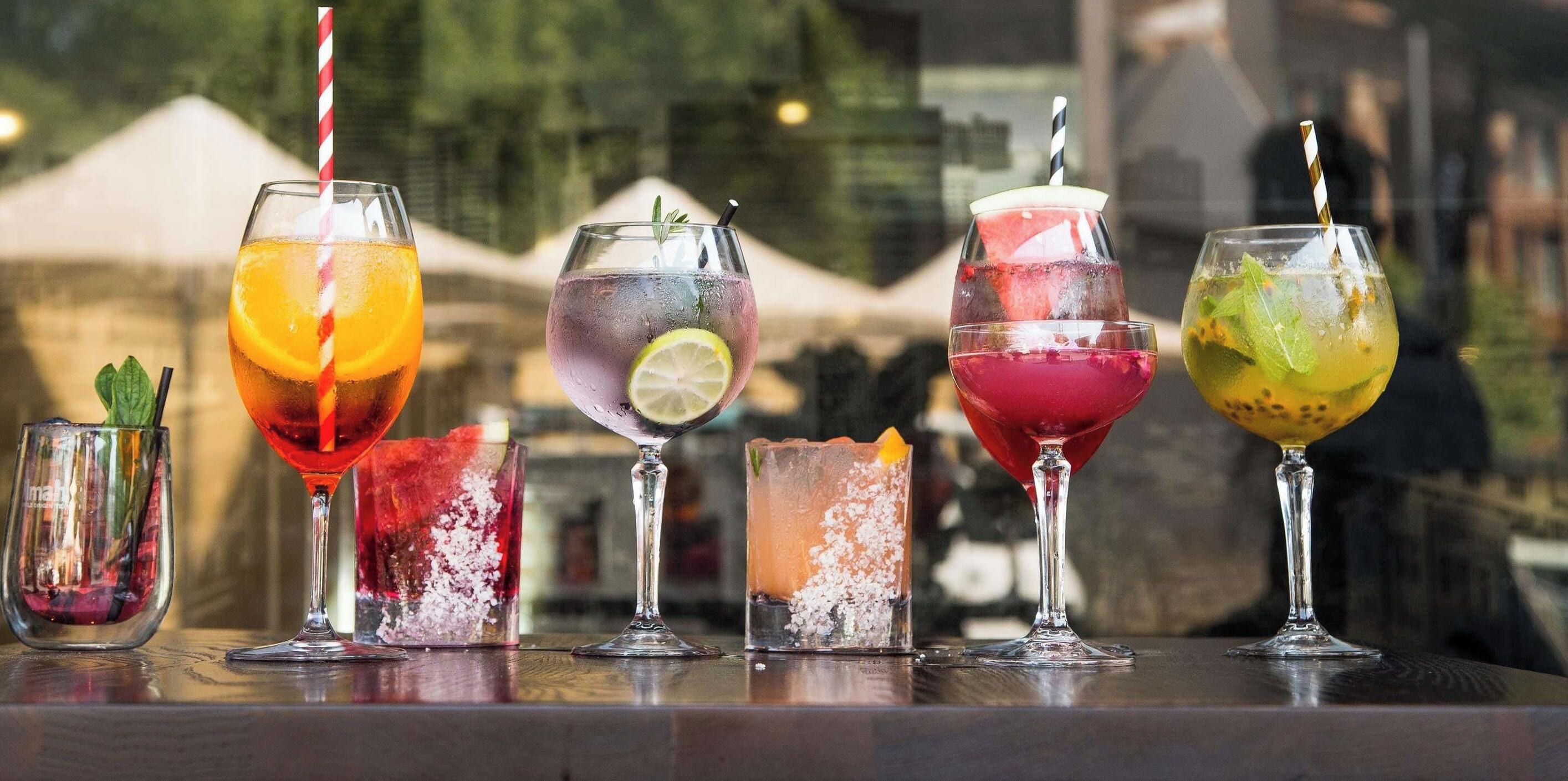 Une variété de cocktails colorés sont présentés sur le comptoir d’un bar, avec une vue sur la ville en arrière-plan.