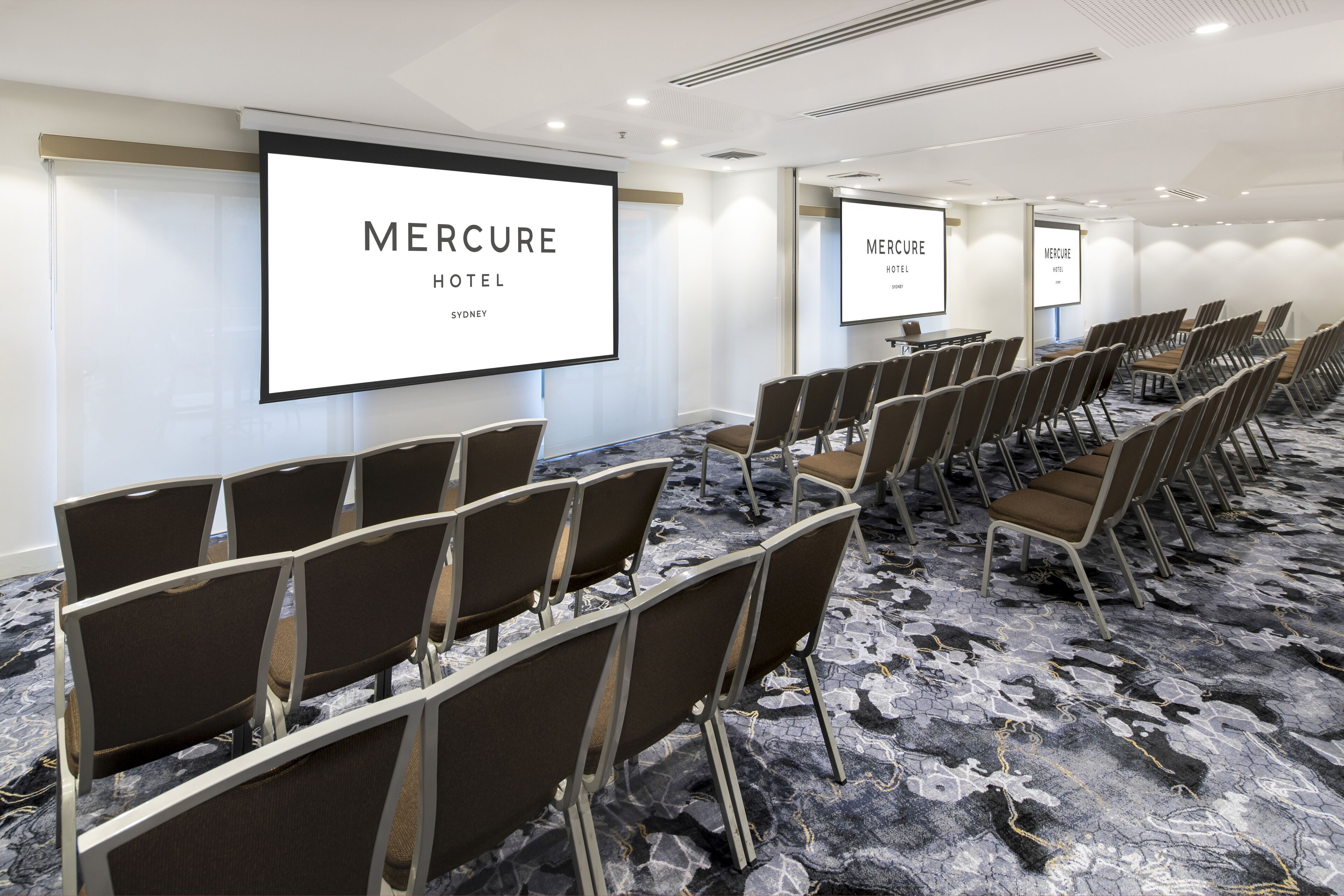 A conference room with rows of chairs, a large screen displaying 'Mercure Hotel Sydney', and a carpeted floor.