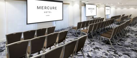 A conference room with rows of chairs, a large screen displaying 'Mercure Hotel Sydney', and a carpeted floor.