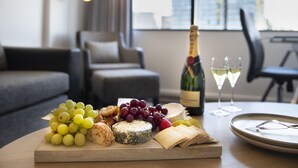 A table with a variety of cheeses, grapes, and a bottle of champagne in a modern living room with a sofa and a window in the background. - Mercure Sydney (Chippendale)