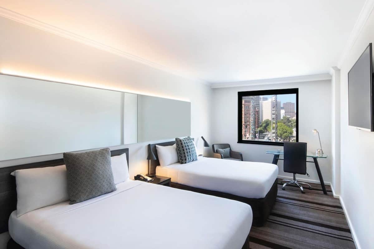 A hotel room with two beds, a desk, a chair, and a large window with a city view.