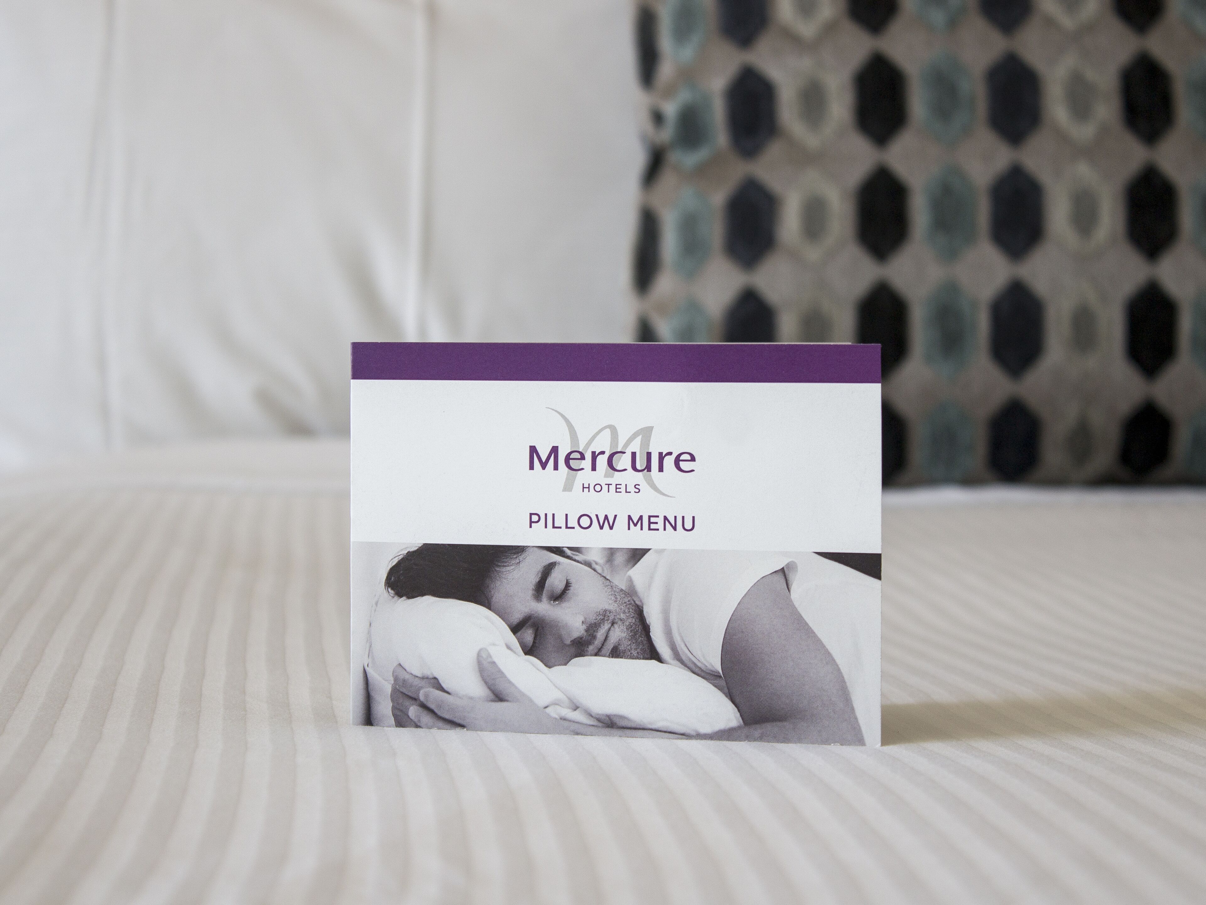 A Mercure Hotels pillow menu on a bed with a patterned pillow in the background.