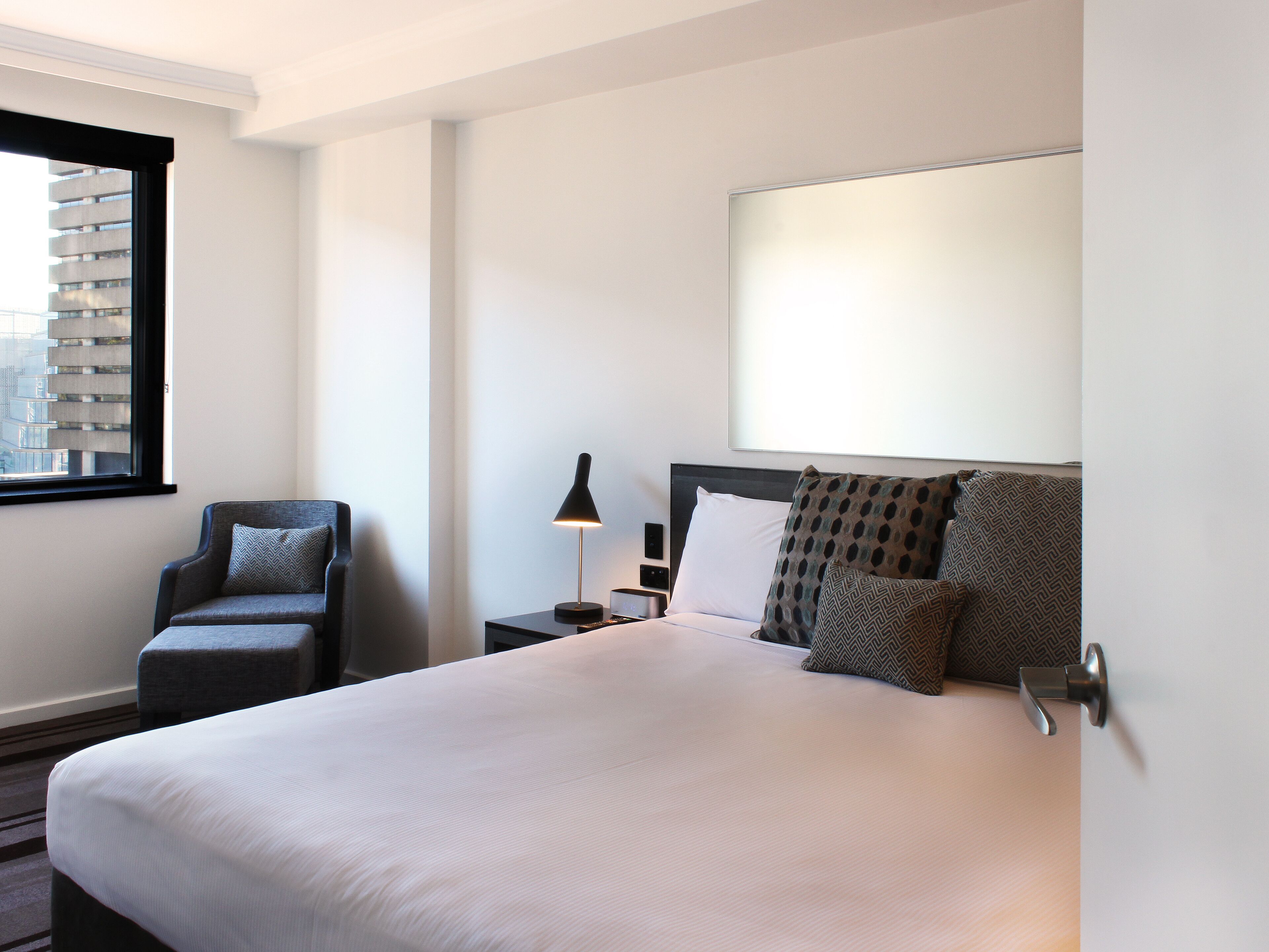 A hotel room with a large bed, a chair, a nightstand, a lamp, and a window with a view of buildings.