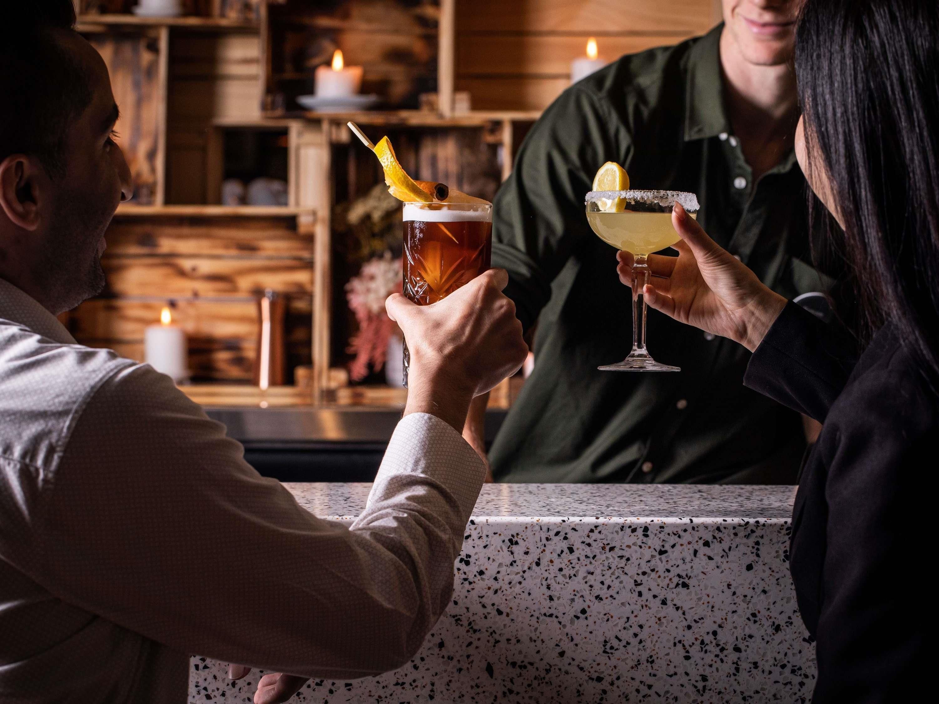 A bar setting with two people holding drinks, a candle, and a wooden background.
