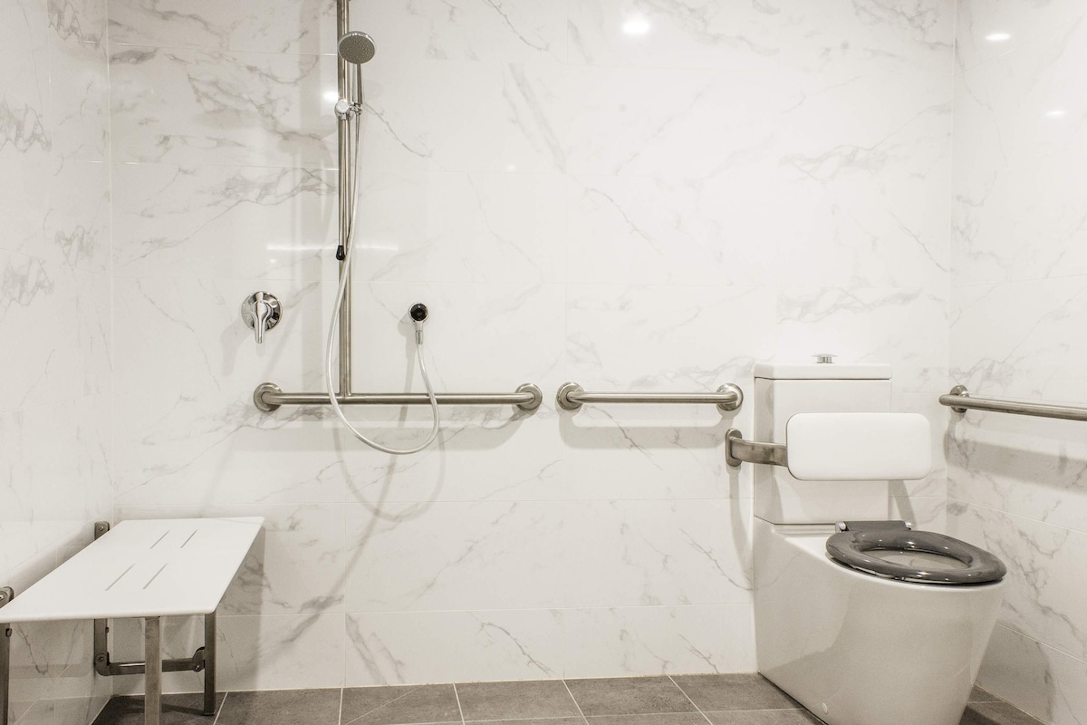 Une salle de bain avec des toilettes blanches, une douche équipée d’un pommeau de douche amovible et d’une barre d’appui.
