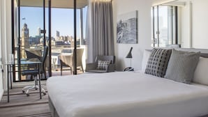 A hotel room with a large bed, a desk with a chair, a city view through floor-to-ceiling windows, and a cityscape painting on the wall. - Mercure Sydney (Chippendale)