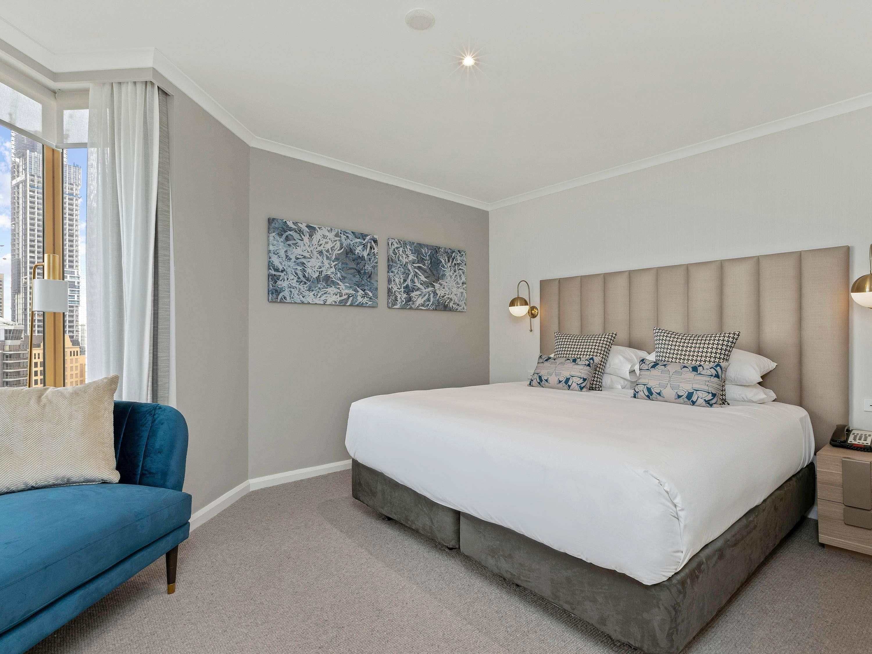 A bedroom with a large bed, a blue sofa, two wall-mounted artworks, and a nightstand with a lamp.