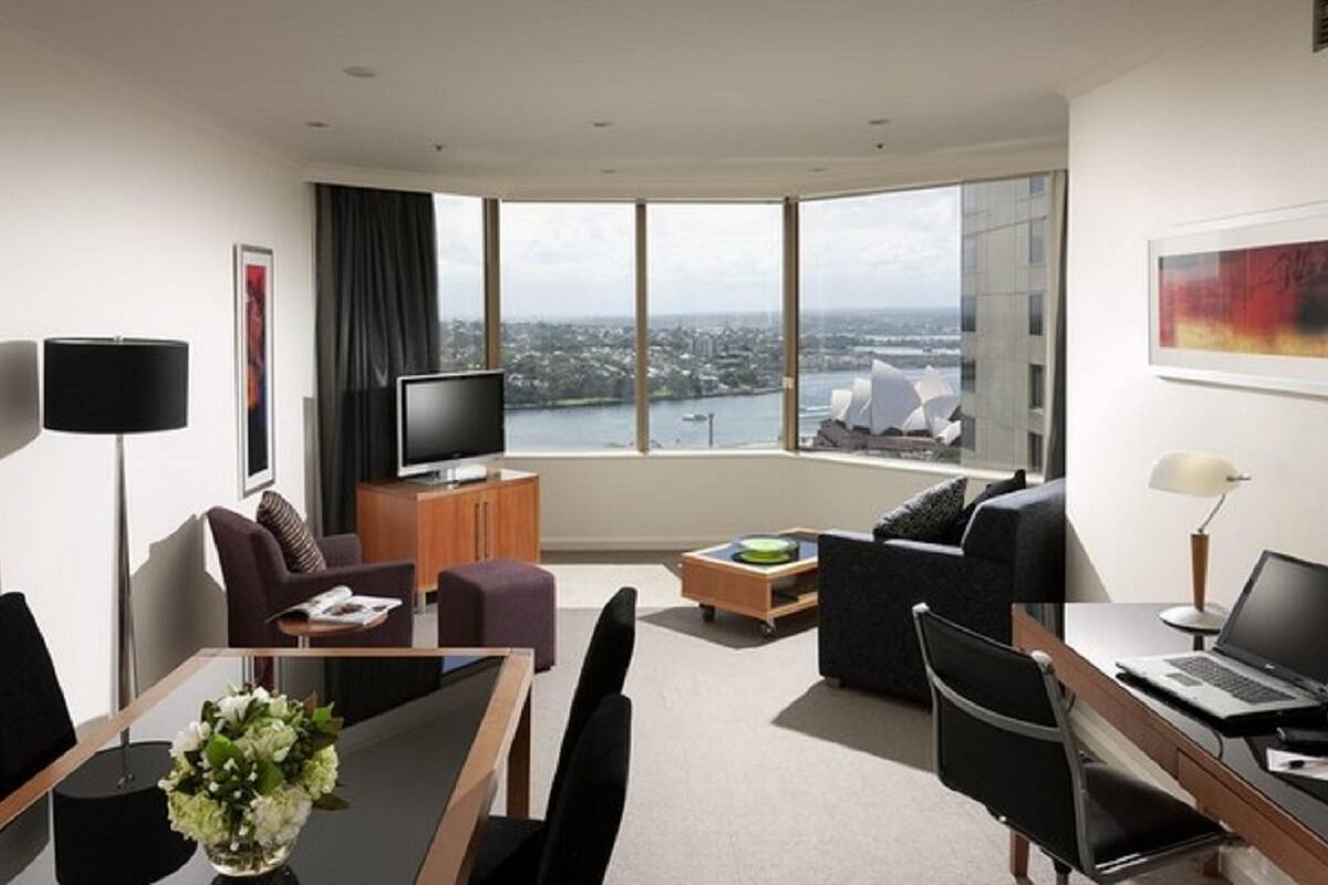 A modern hotel room with a large window offering a city view, a flat-screen TV, a dining area with a table and chairs, and a seating area with a sofa and armchairs.