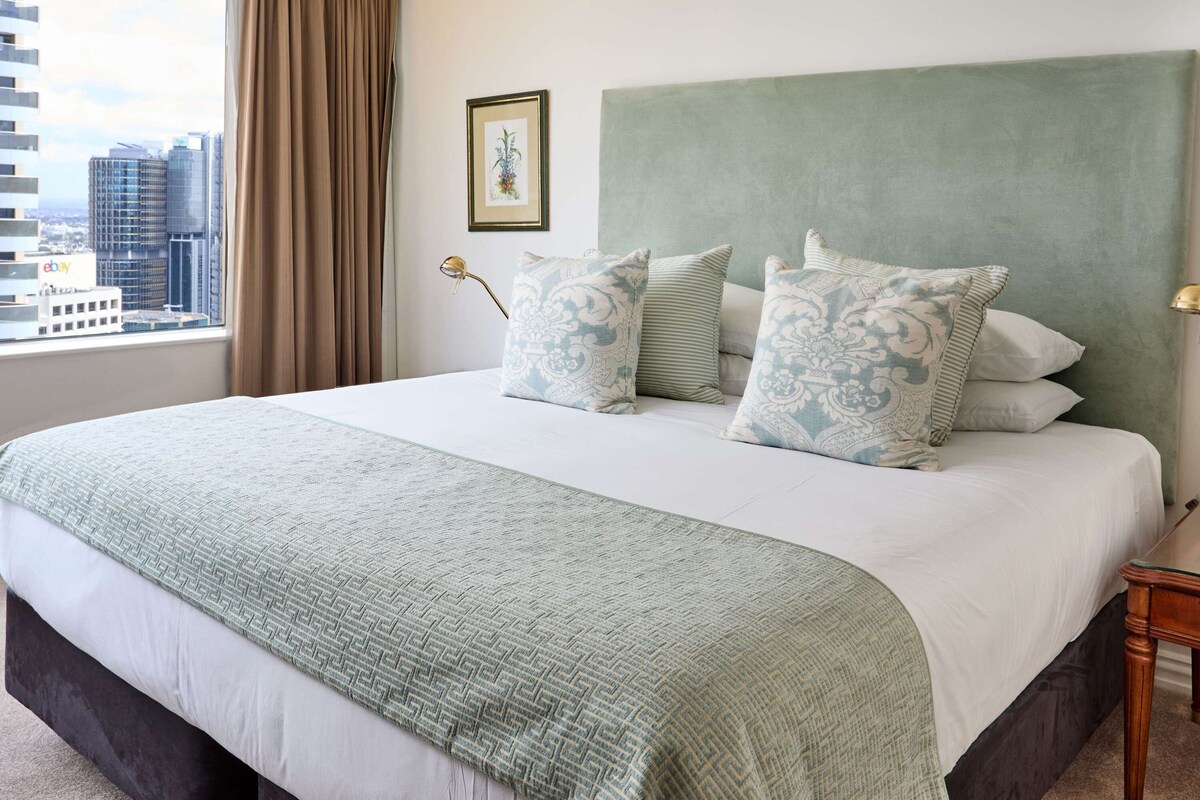 A hotel room with a large bed, a view of the city, and a framed picture on the wall.