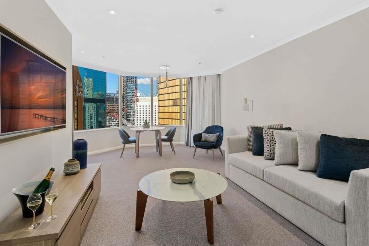 A modern living room with a sofa, a coffee table, a dining table with chairs, and a large window with a city view.