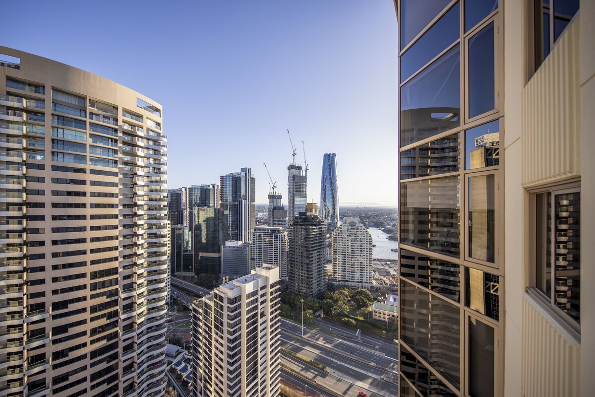 A high-rise building with multiple floors and balconies, surrounded by other tall buildings and a river.
