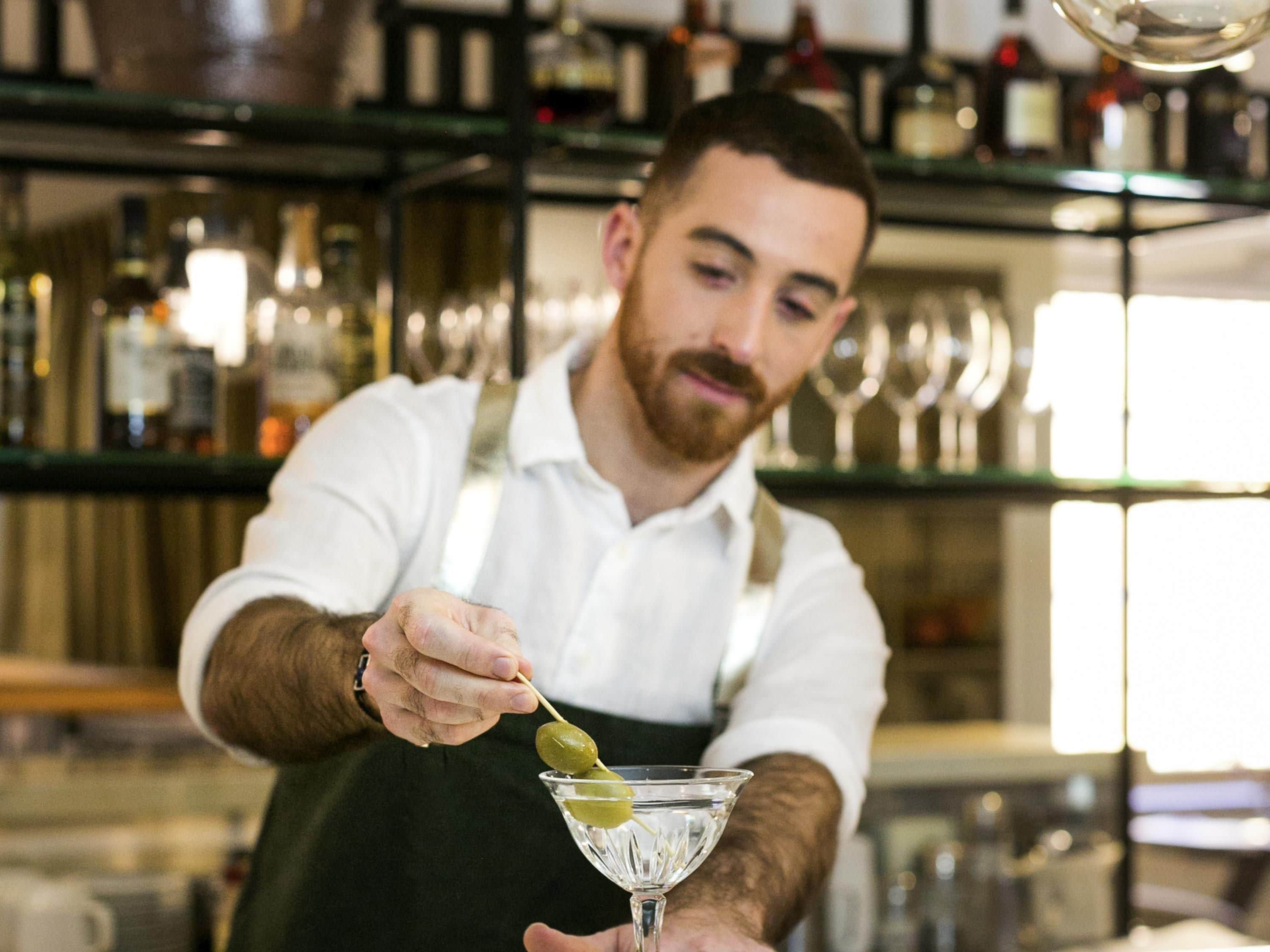 Un barman prépare un cocktail dans un bar, avec des étagères remplies de bouteilles et de verres en arrière-plan.