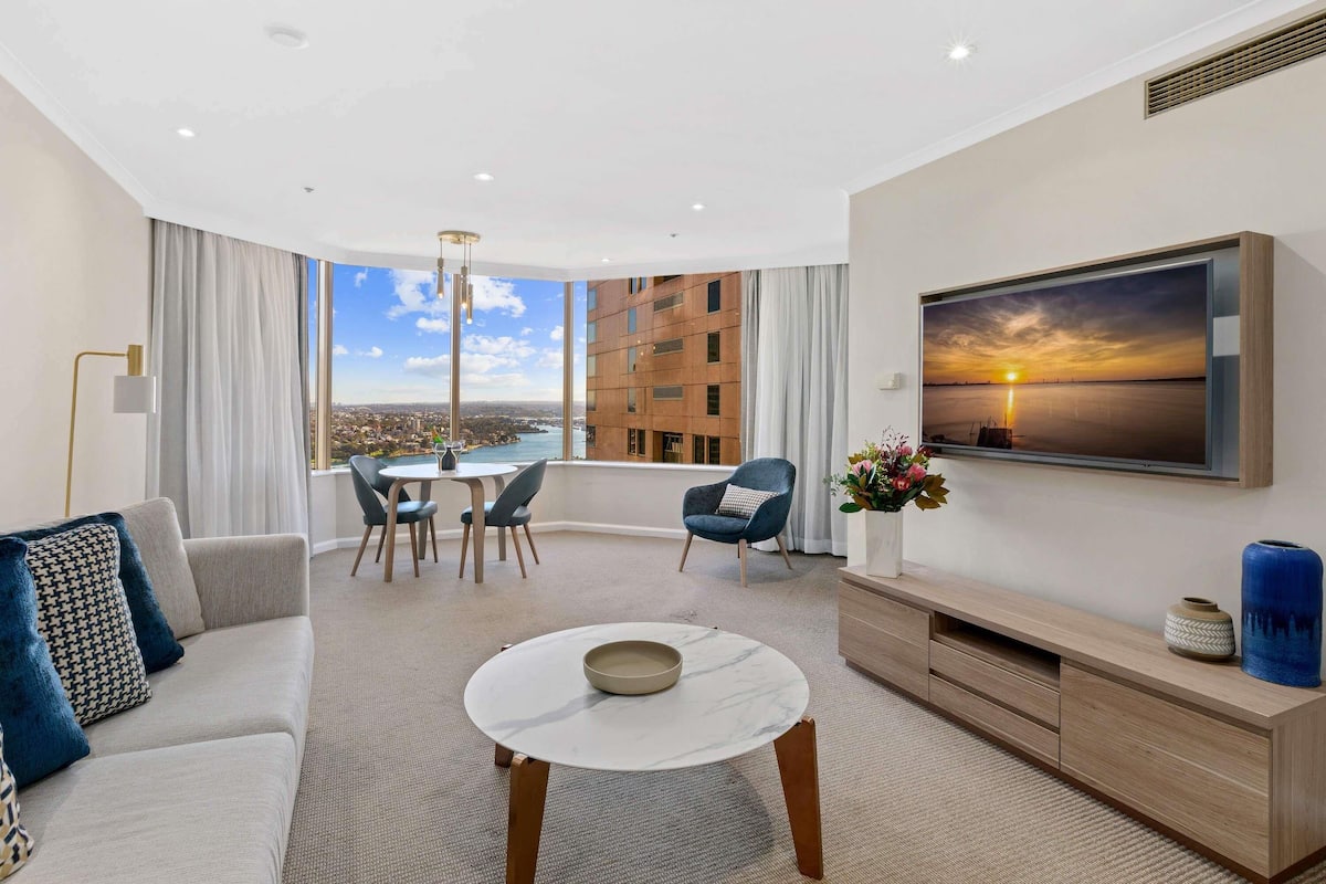 A modern living room with a sofa, a coffee table, a dining area with chairs, a TV mounted on the wall, and a large window offering a city view.