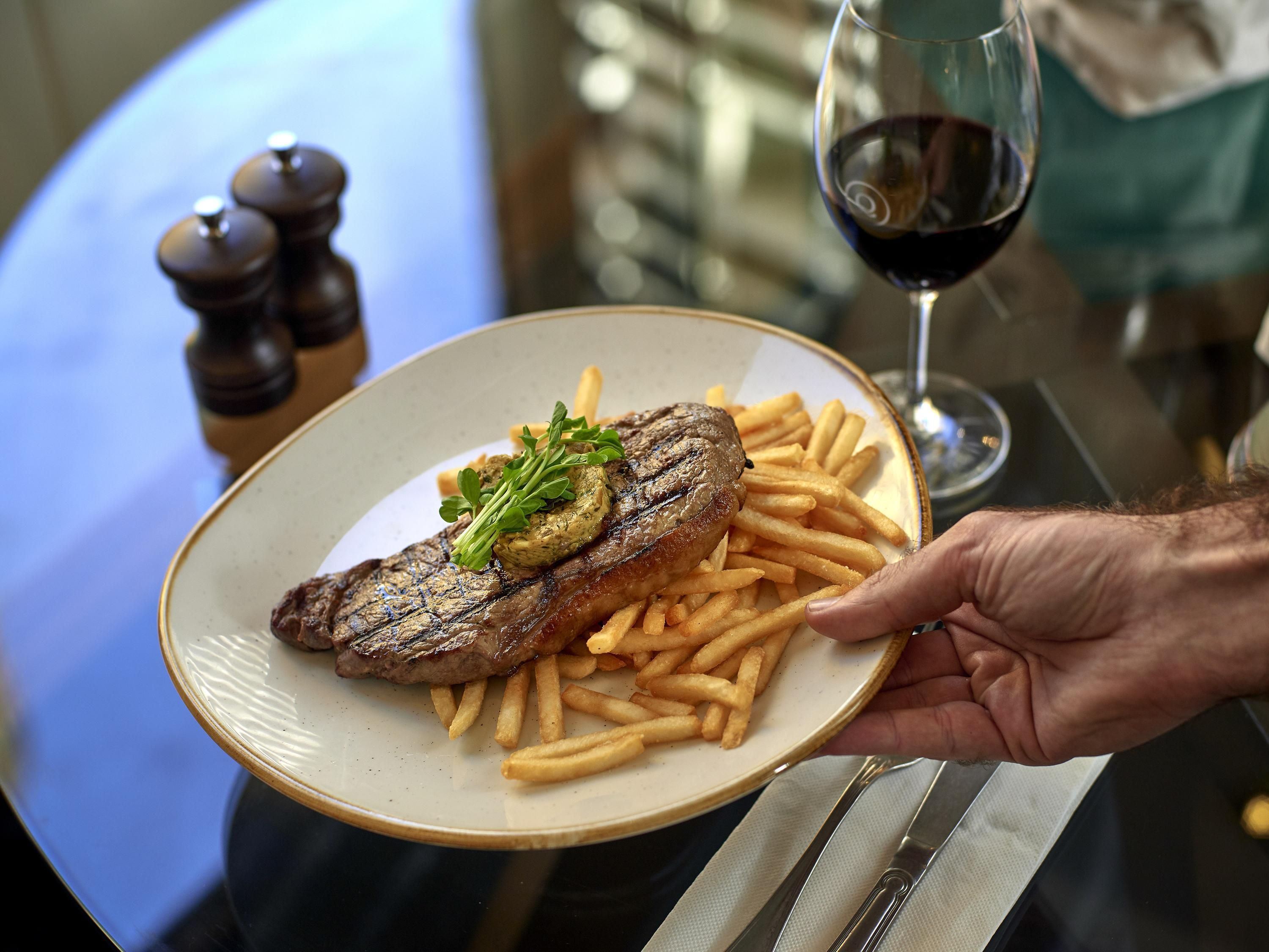 A plate with a steak and fries, a glass of red wine, and a hand holding the plate.