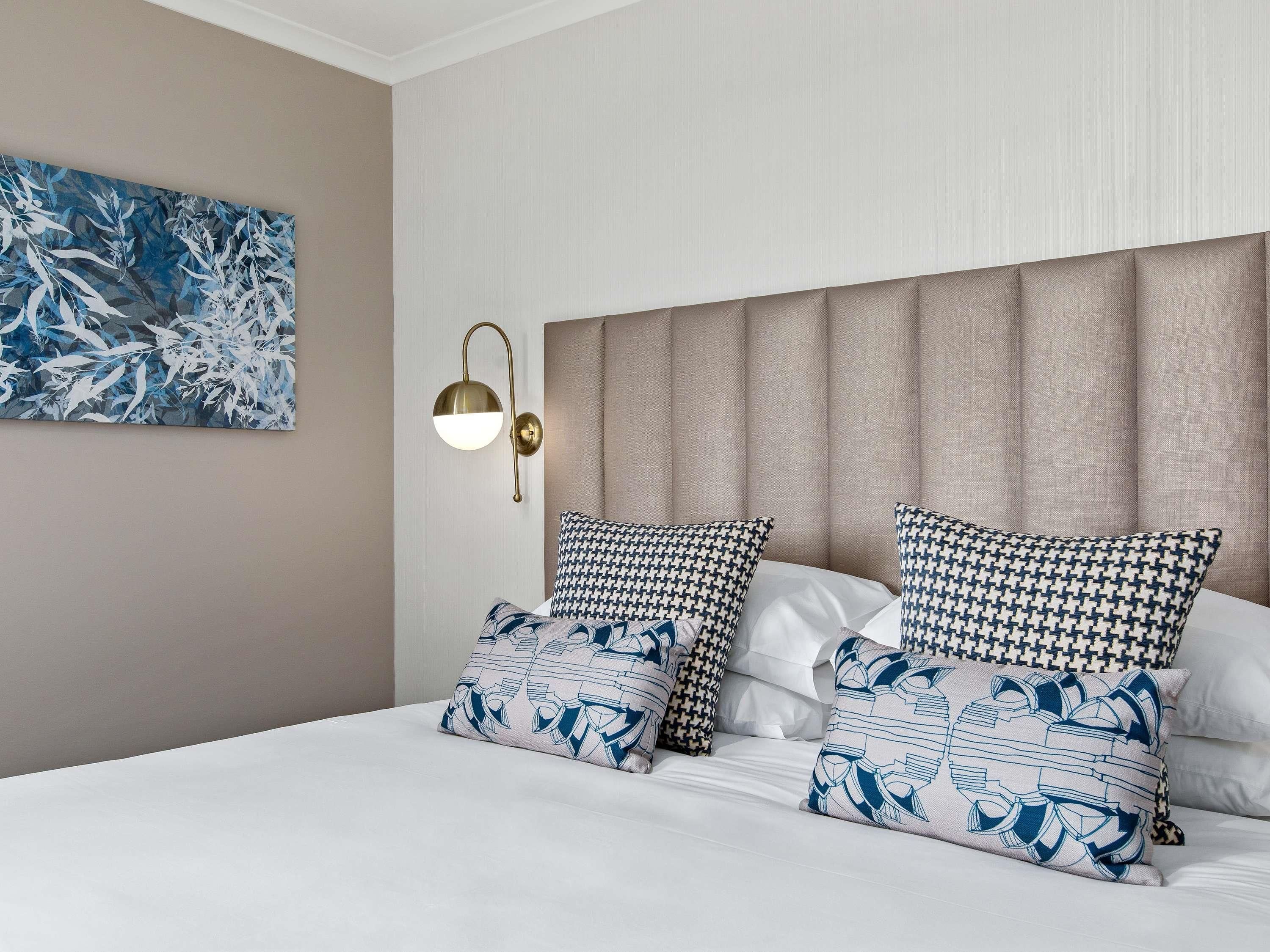 A hotel room with a bed, two patterned pillows, a wall-mounted light fixture, and a large abstract painting.