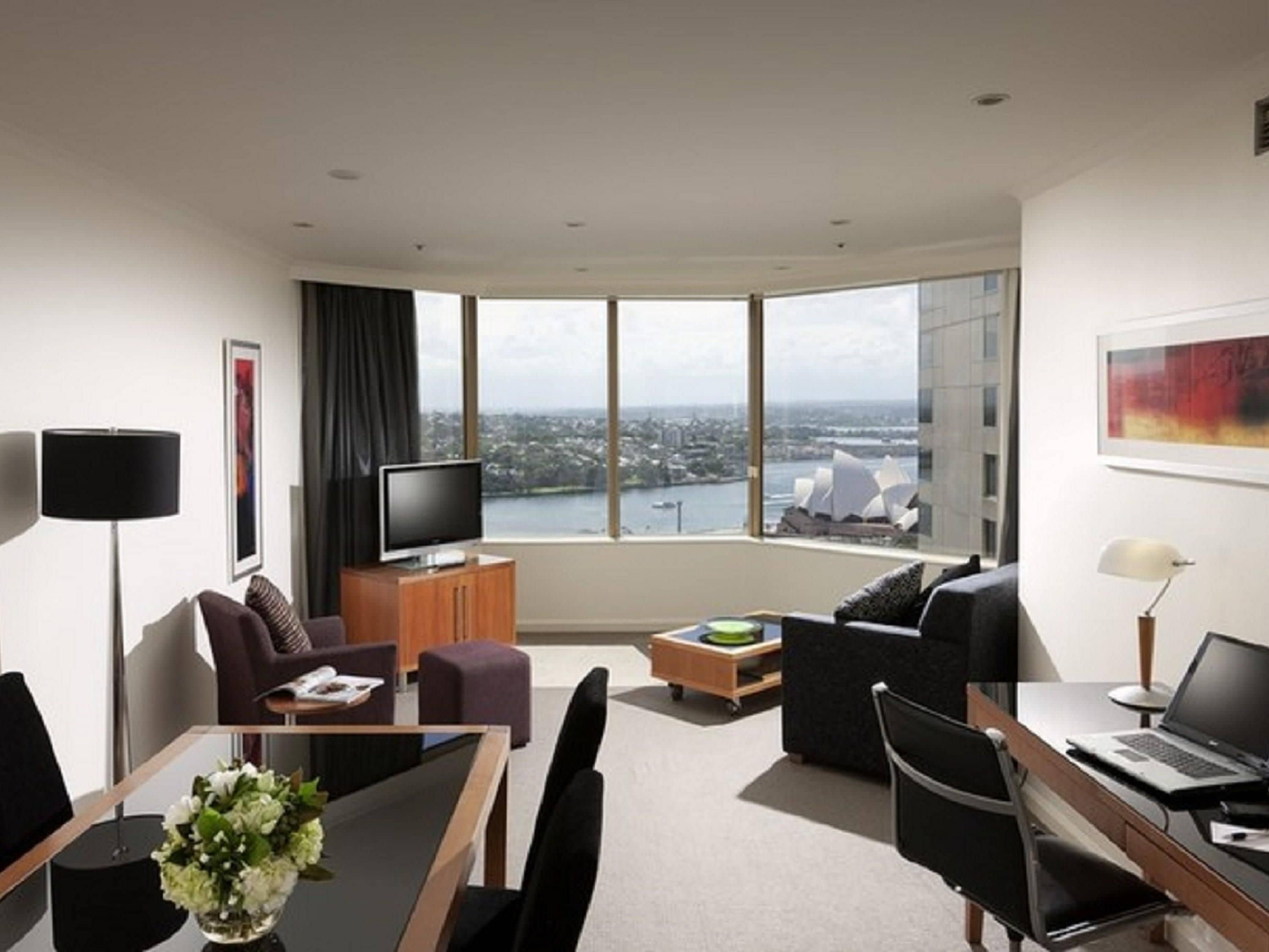 Habitación de hotel moderna con un ventanal grande que ofrece vistas a la ciudad, un televisor de pantalla plana, una zona de comedor con mesa y sillas, y un área de descanso con sofá y sillones.