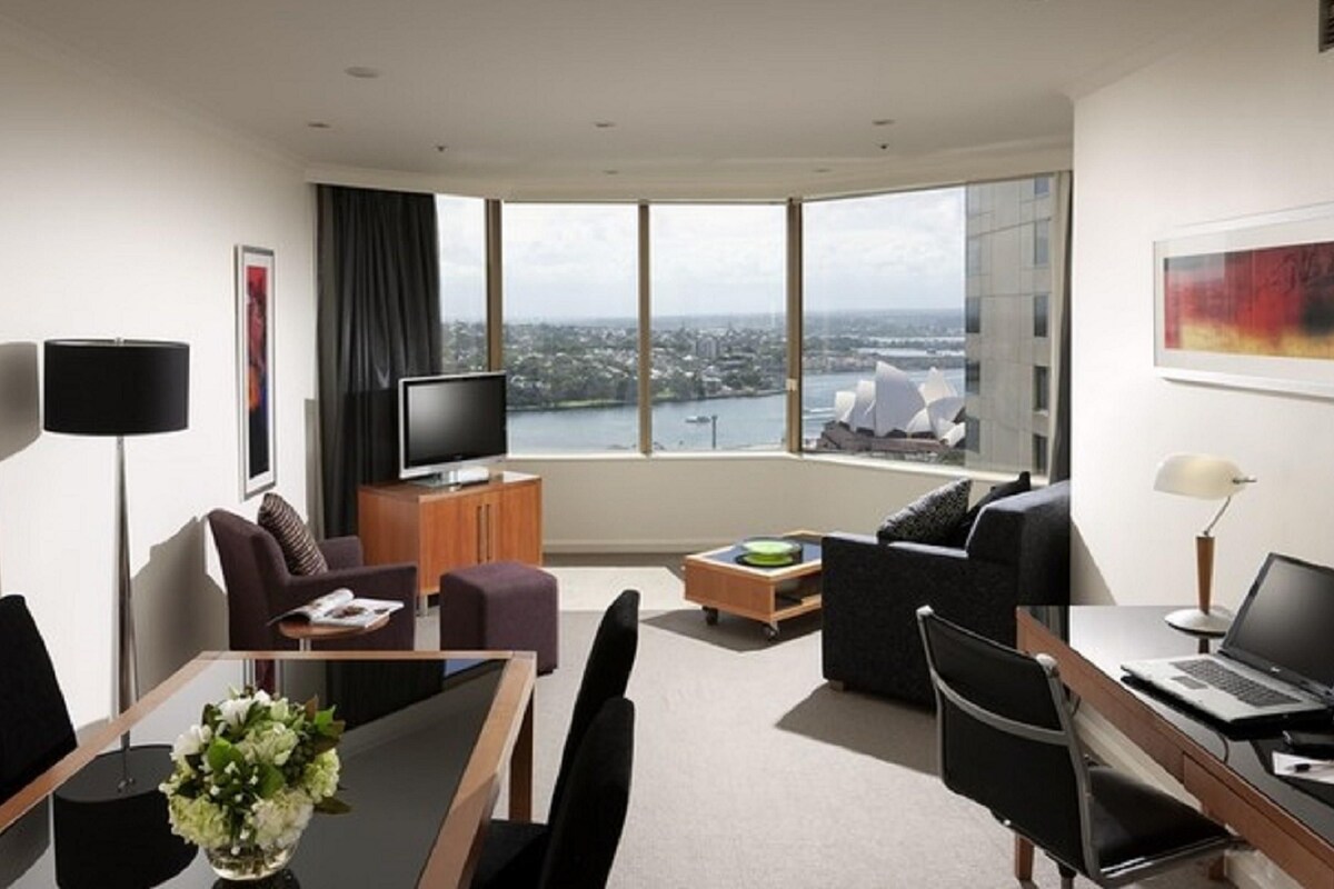 A modern hotel room with a large window offering a city view, a flat-screen TV, a dining area with a table and chairs, and a seating area with a sofa and armchairs.