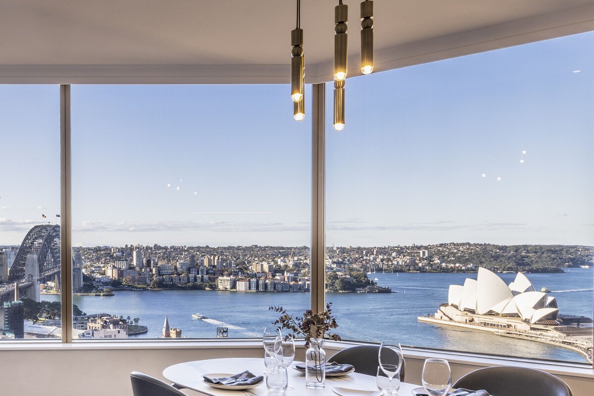 A dining area with a table set for four, large windows offering a view of a city skyline and a famous opera house.