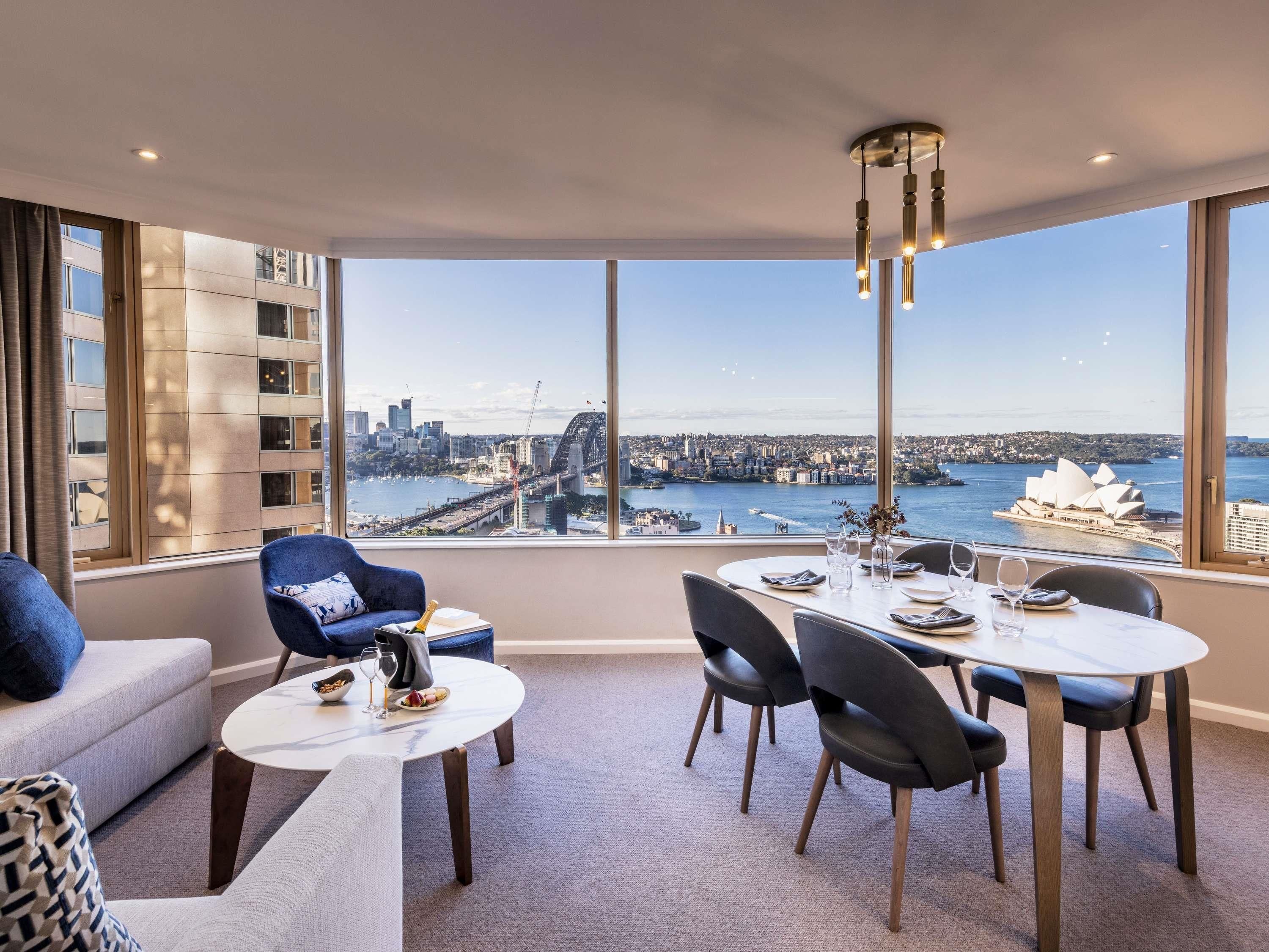 A dining area with a table set for four, a sofa, and a view of a cityscape with a bridge and the Sydney Opera House.