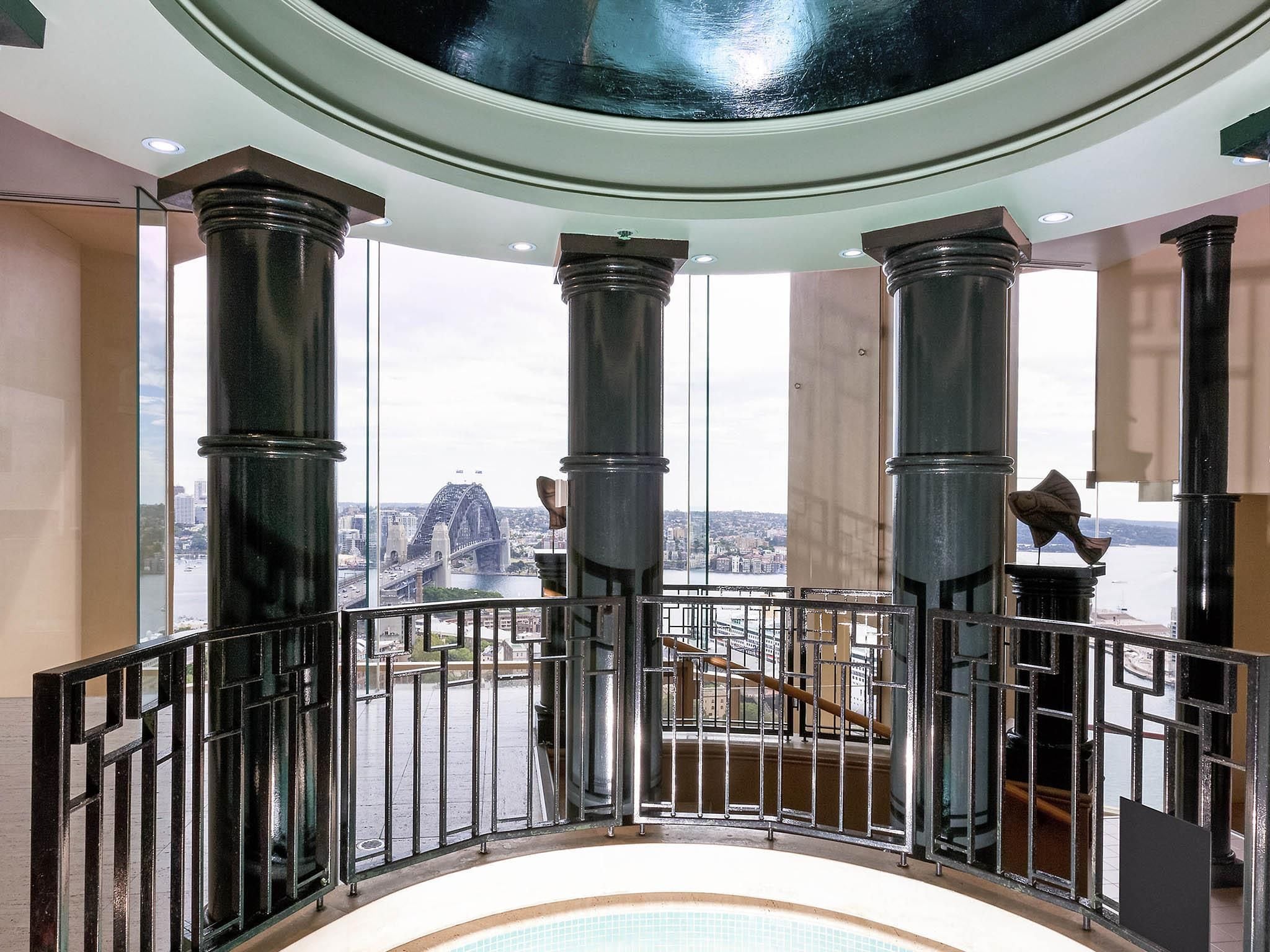 Un balcon offrant une vue sur le pont du port de Sydney, avec de grandes colonnes et une balustrade en verre.