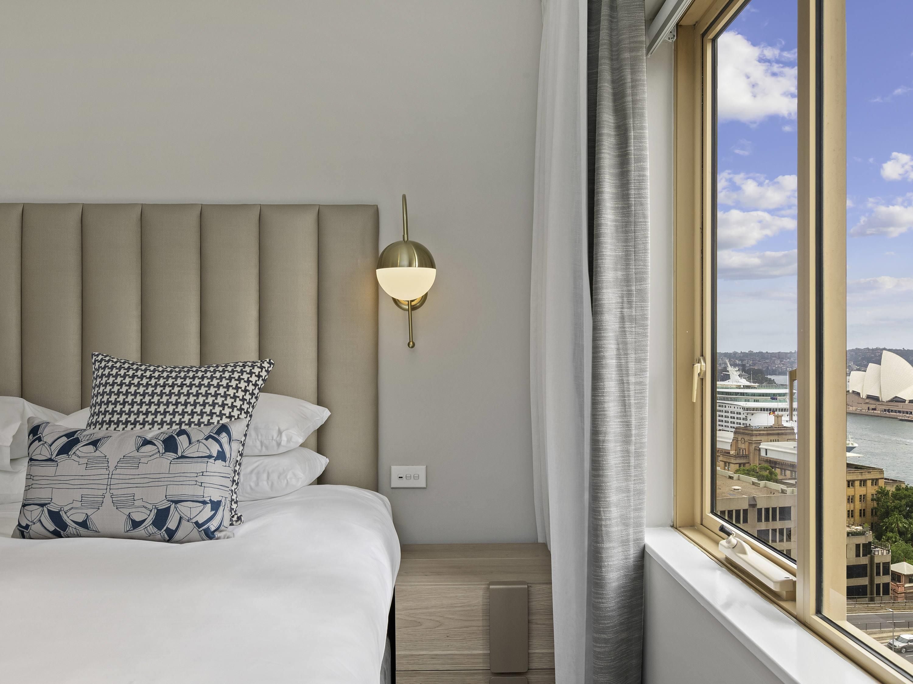 Habitación de hotel con una cama grande, una lámpara de pared y una ventana con vistas a un paisaje urbano y un famoso teatro de ópera.