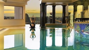 A spacious indoor pool area with a clear view of the sunset over the water, large columns, and a tiled wall with a fish pattern. - The Sebel Quay West Suites Sydney (The Rocks)