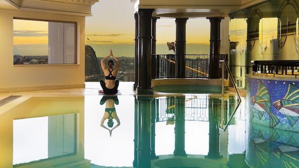 A spacious indoor pool area with a clear view of the sunset over the water, large columns, and a tiled wall with a fish pattern. - The Sebel Quay West Suites Sydney (The Rocks)