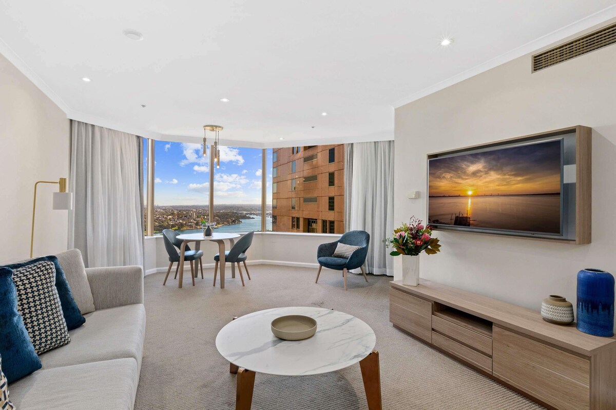 A modern living room with a sofa, a coffee table, a dining area with chairs, a TV mounted on the wall, and a large window offering a city view.