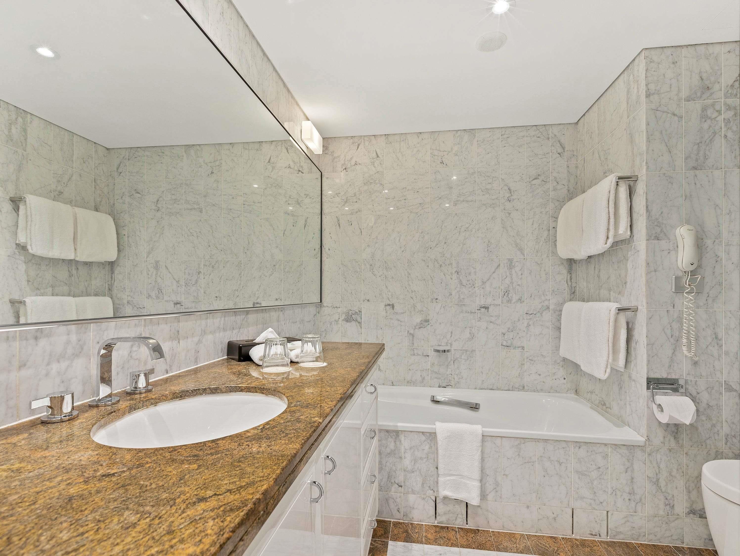 Une salle de bain moderne avec un lavabo surmonté d’un plan de travail en granit, un grand miroir, une baignoire et des toilettes.