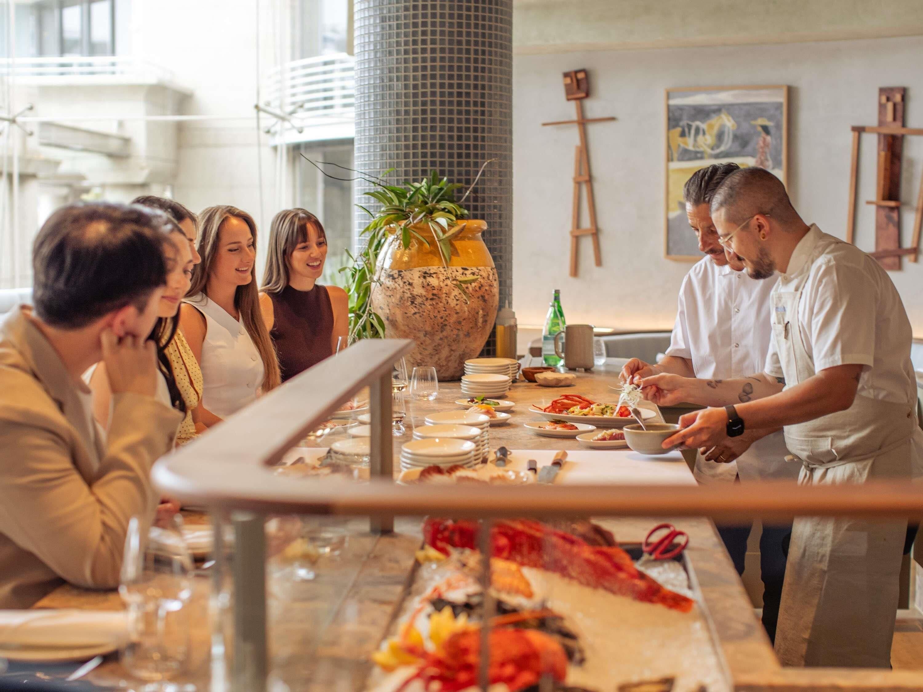 Een groep mensen dineert in een restaurant met een grote vaas en een schilderij aan de muur.