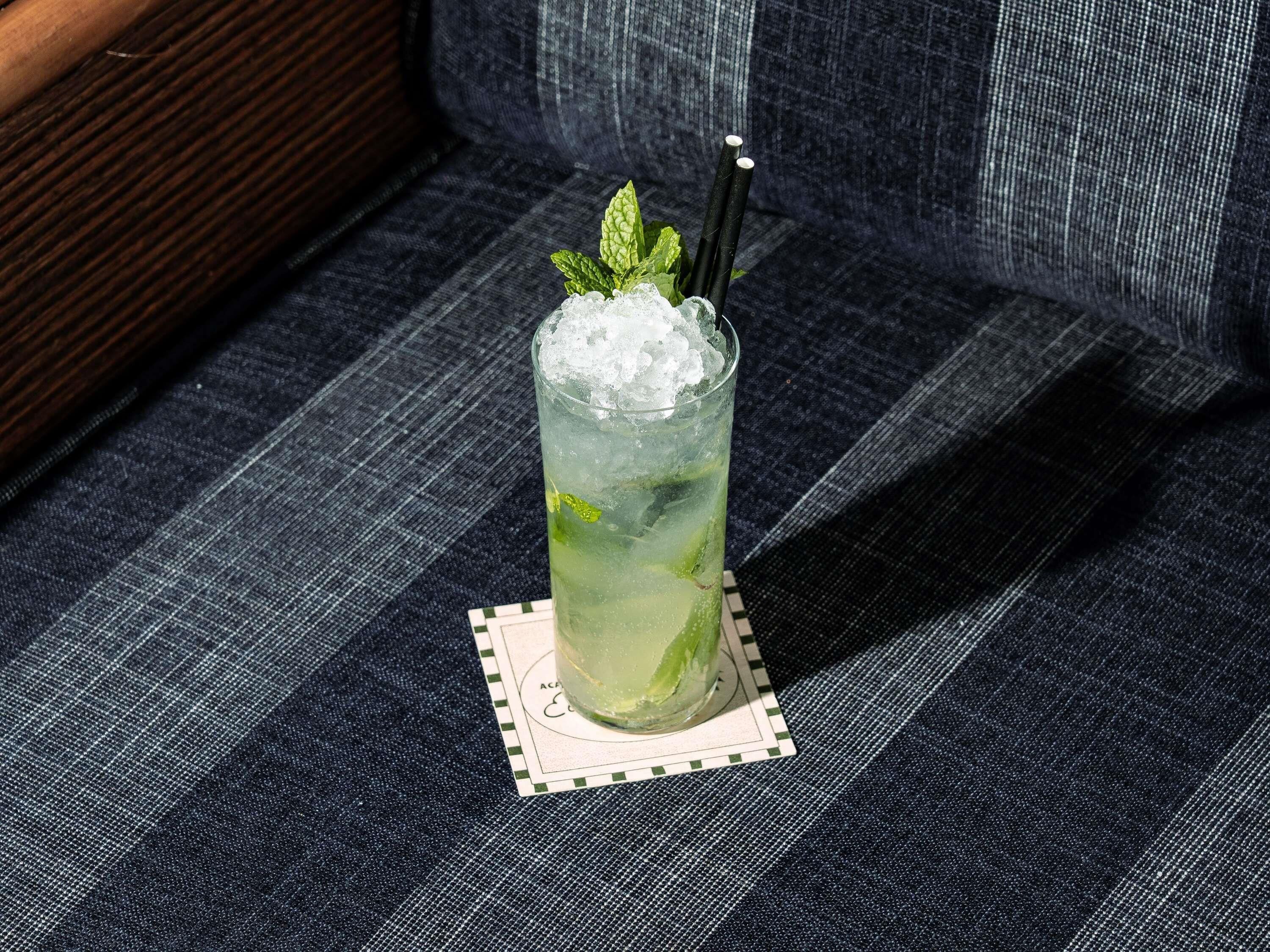 A glass of iced beverage with mint leaves on a coaster, placed on a textured fabric surface.