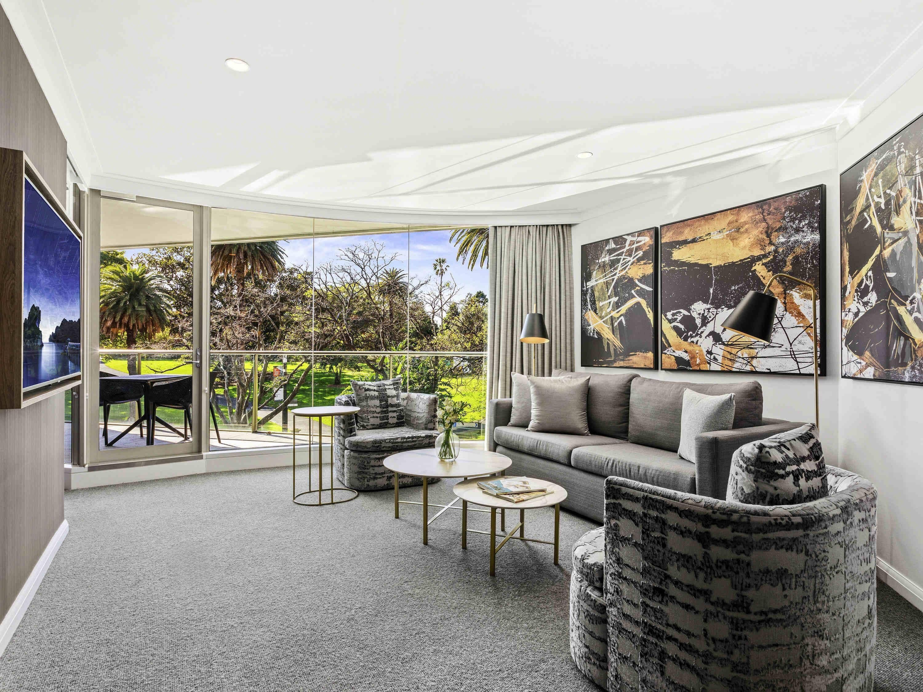 Ein modernes Wohnzimmer mit einem grauen Sofa, zwei Sesseln, einem Couchtisch und einem Fernseher. An den Wänden befinden sich abstrakte Gemälde und ein Balkon mit Blick auf Grünflächen und ein Gewässer.