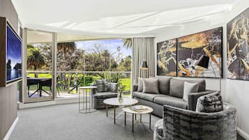 Ein modernes Wohnzimmer mit einem grauen Sofa, zwei Sesseln, einem Couchtisch und einem Fernseher. An den Wänden befinden sich abstrakte Gemälde und ein Balkon mit Blick auf Grünflächen und ein Gewässer.