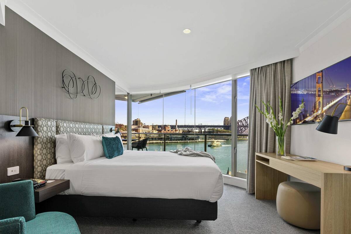 A hotel room with a large bed, a desk, a chair, a view of a bridge, and a cityscape.