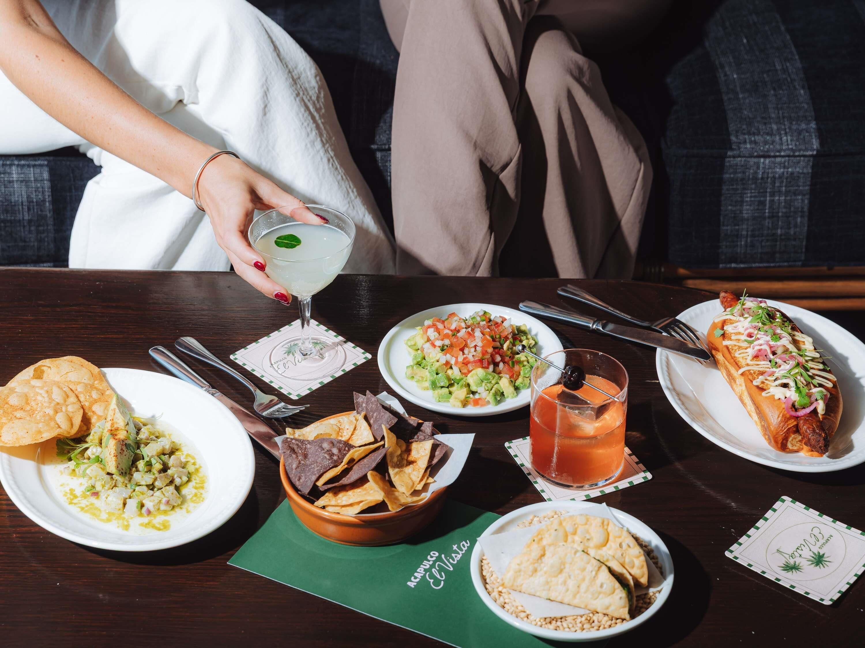 A dining table with various dishes, a cocktail, and a person holding a drink.