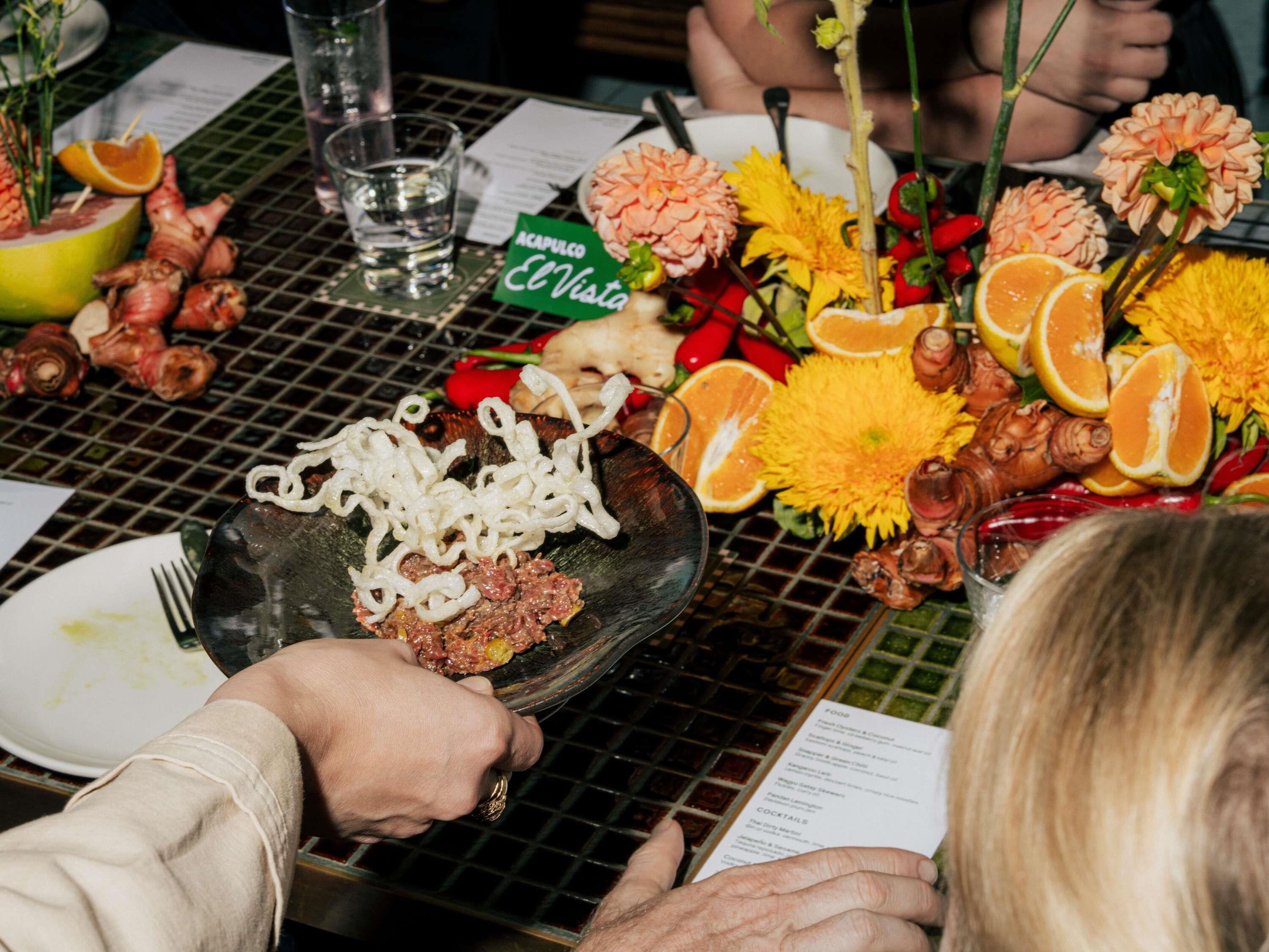 Une table à manger sur laquelle sont disposés divers aliments, notamment un plat de nouilles et de viande, le tout entouré de fruits et de fleurs.