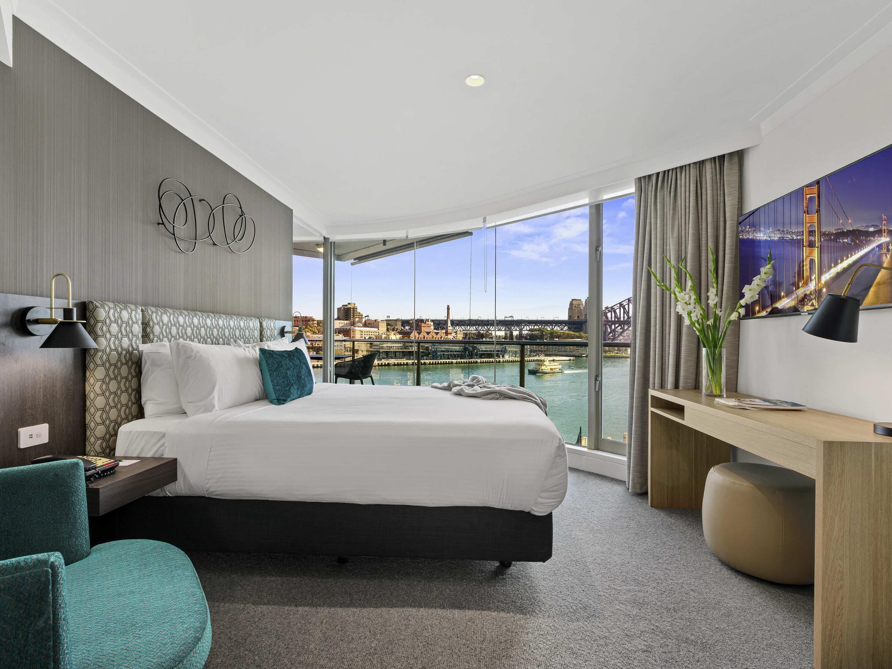 A hotel room with a large bed, a desk, a chair, a view of a bridge, and a cityscape.