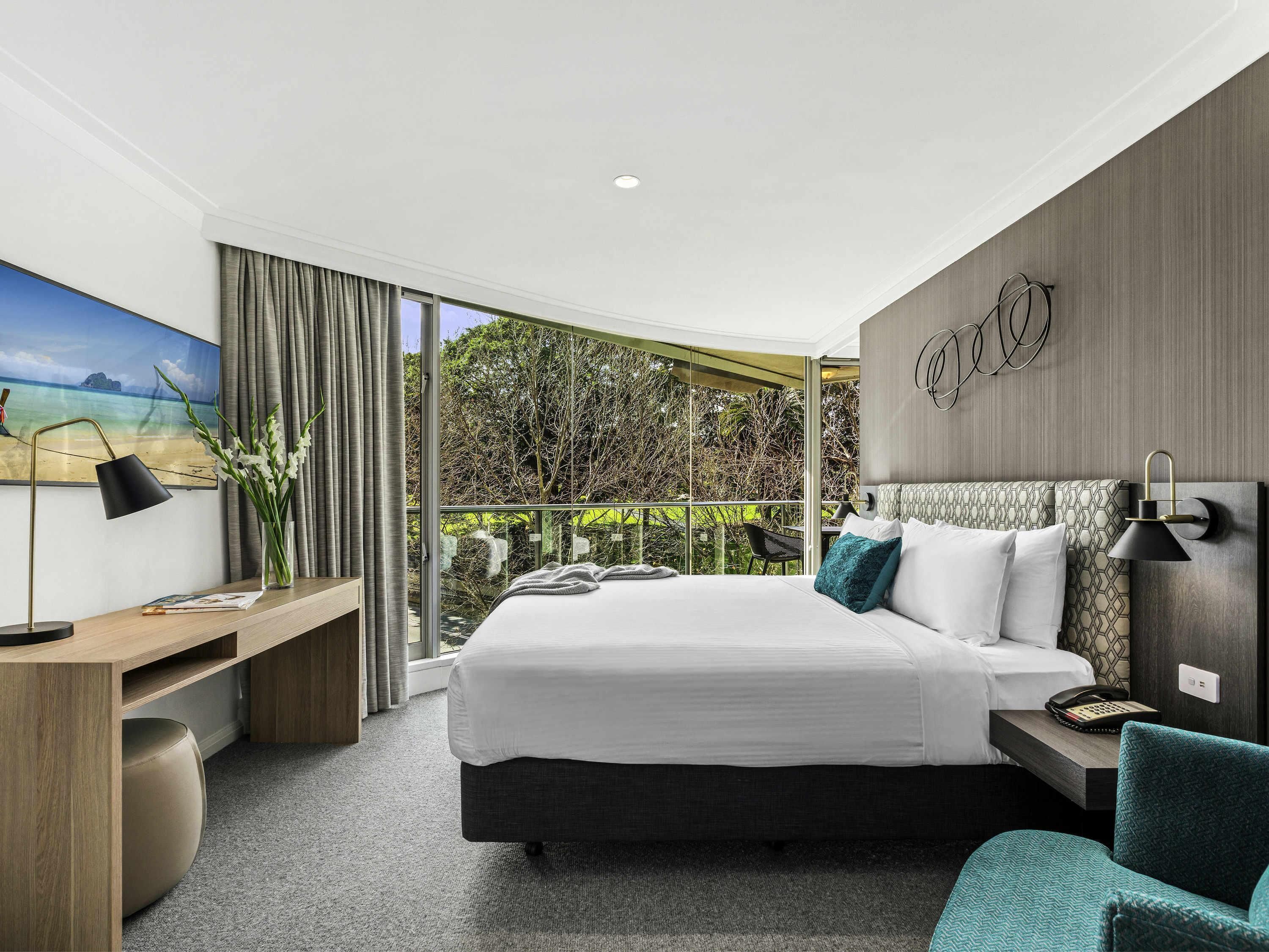 A modern hotel room with a large bed, a desk, a chair, and a balcony with a view of greenery.