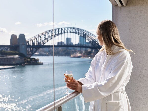 A person in a white robe holding a glass of drink, overlooking a body of water with a bridge in the background. - Pullman Quay Grand Sydney Harbour (Sydney)