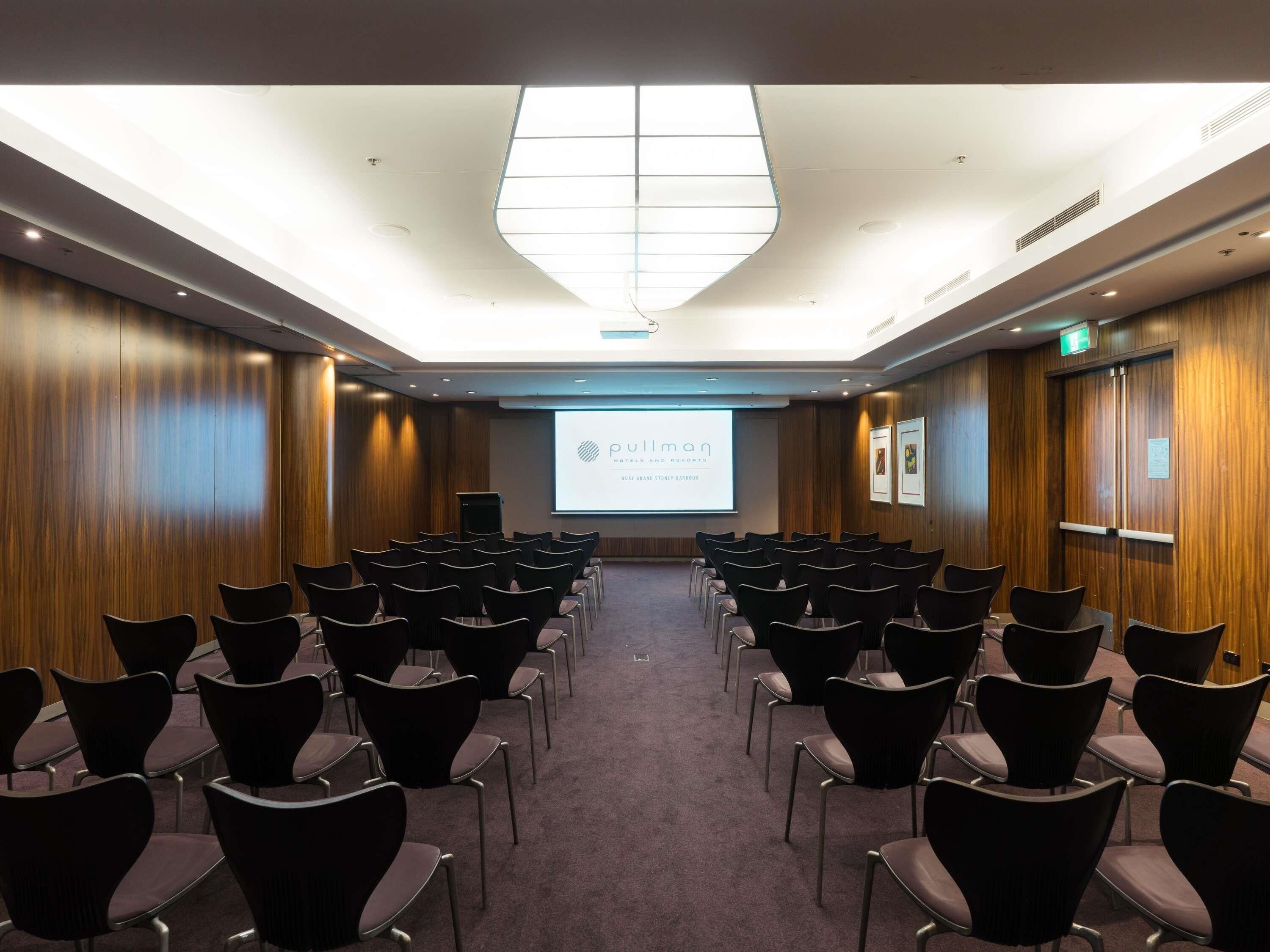 Une salle de conférence avec des rangées de chaises disposées face à un écran de projection sur lequel est affiché un diaporama.