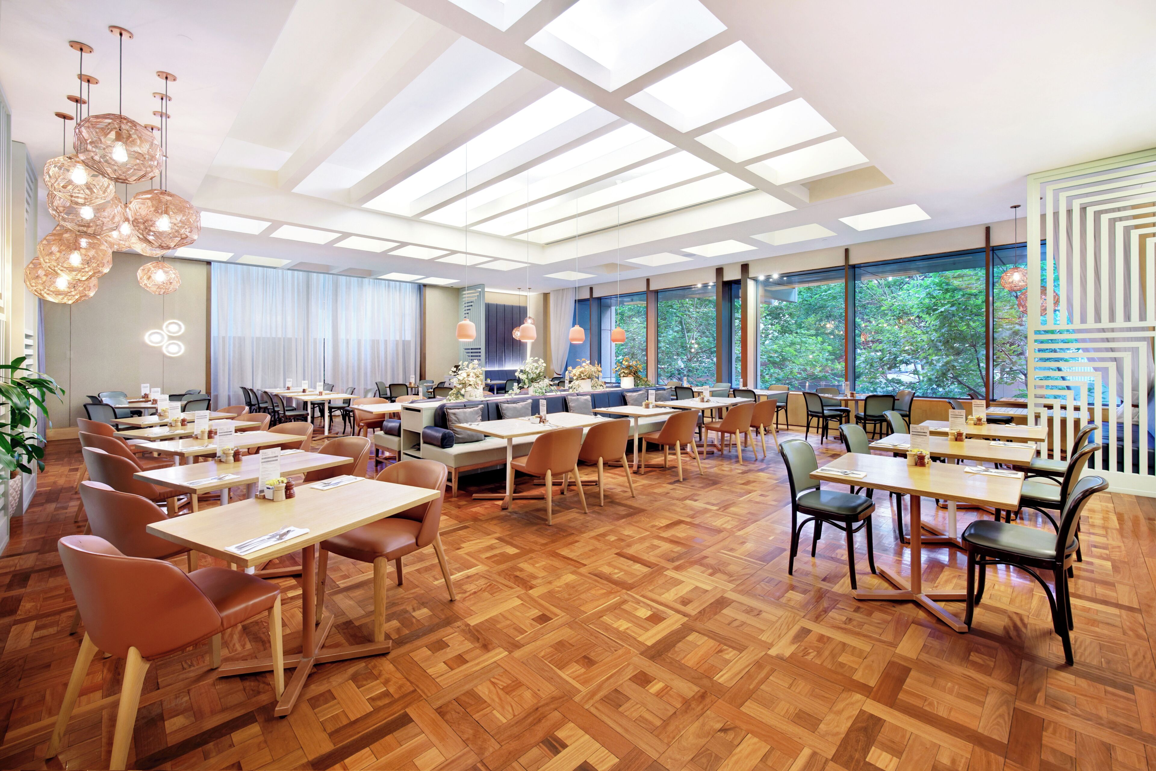 A spacious dining area with wooden flooring, multiple tables and chairs, and large windows offering a view of the outdoors.
