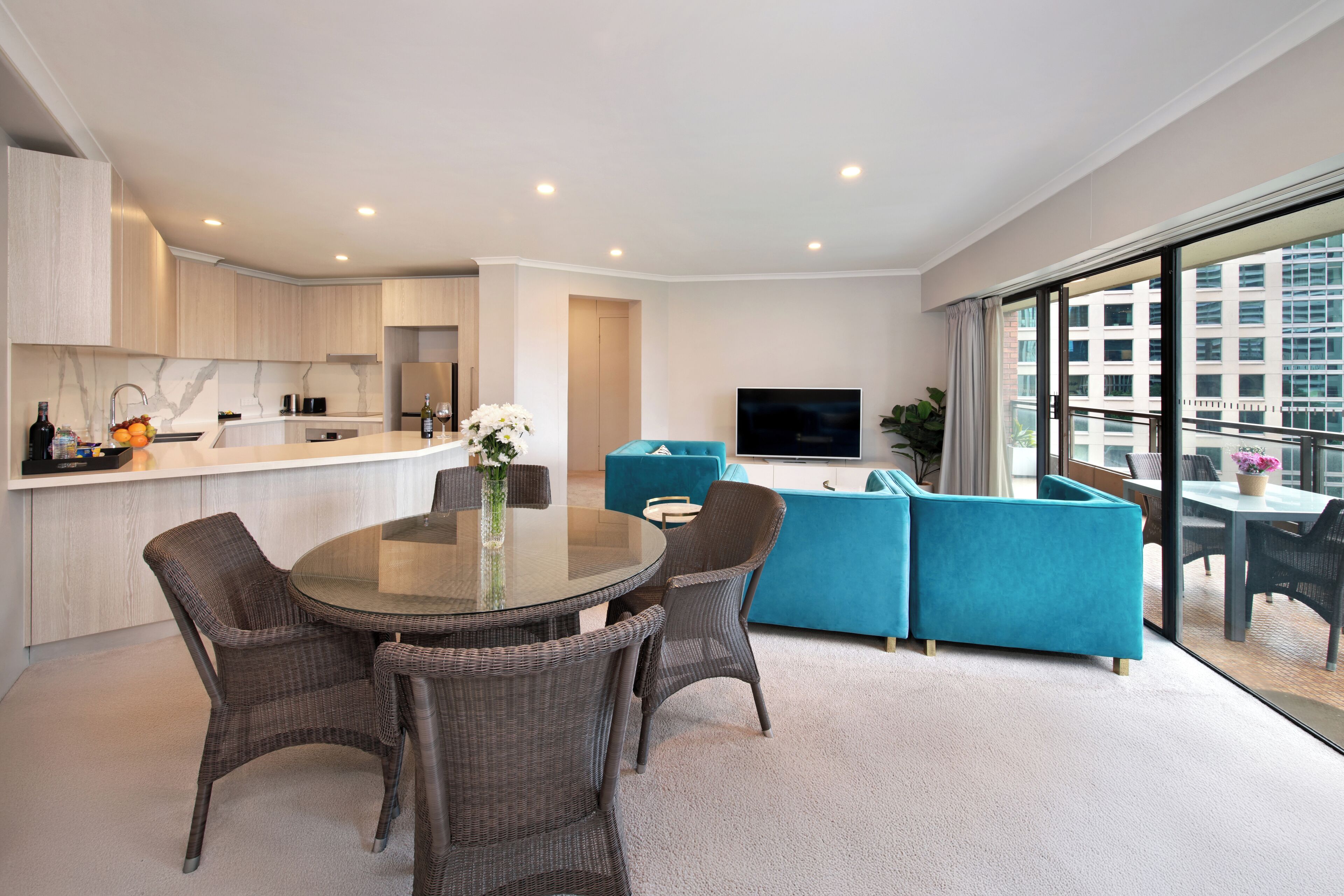A modern apartment with a kitchen, dining area, and living room. It features a large window, a flat-screen TV, and a balcony with a view of the city.