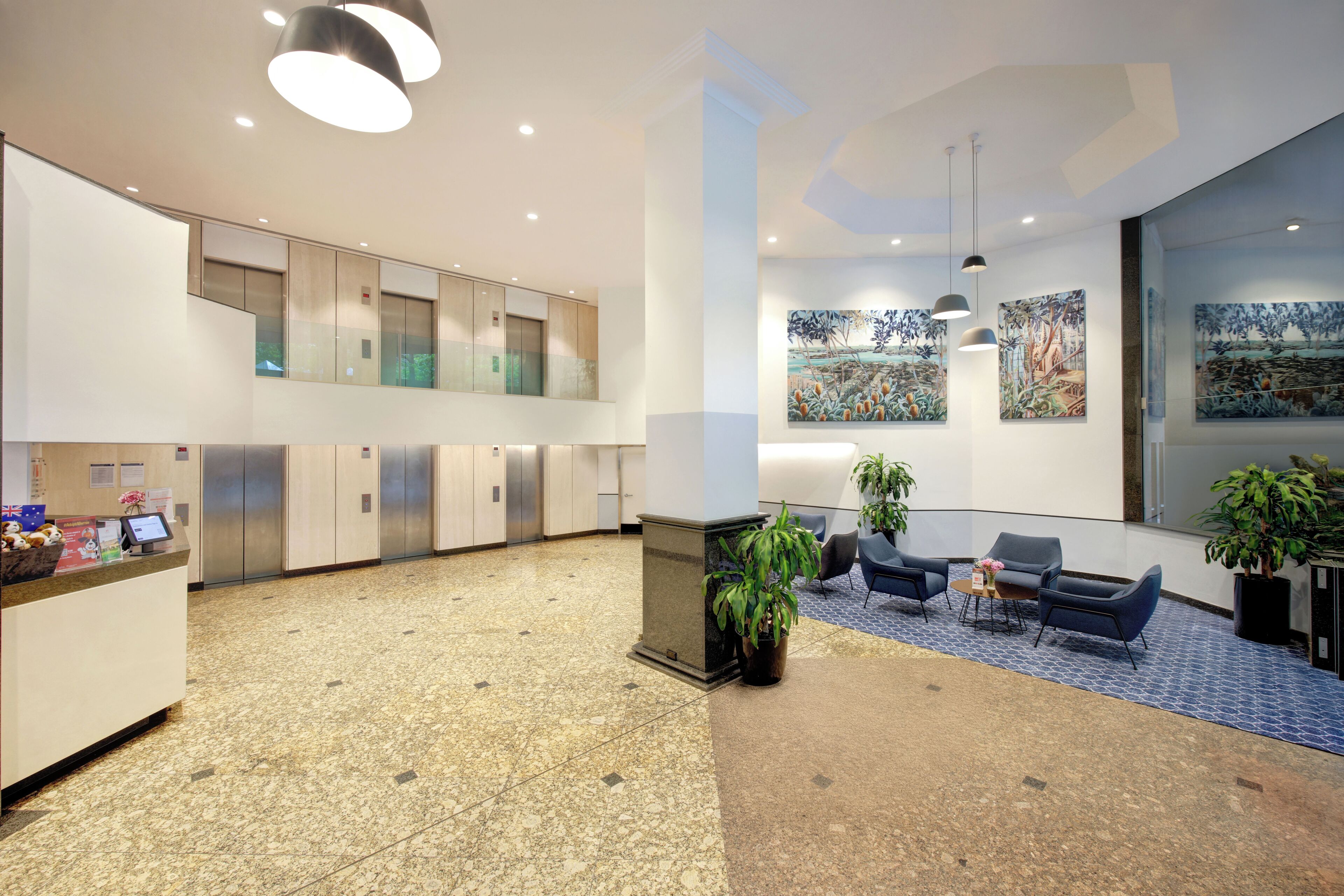 A modern lobby with a reception desk, seating area, artwork on the walls, and a staircase leading to an upper level.