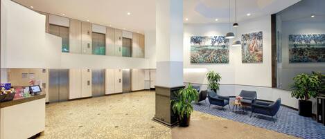 A modern lobby with a reception desk, seating area, artwork on the walls, and a staircase leading to an upper level.