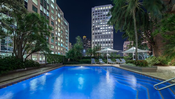 Una piscina con sillones de playa y sombrillas, rodeada de edificios altos y palmeras.