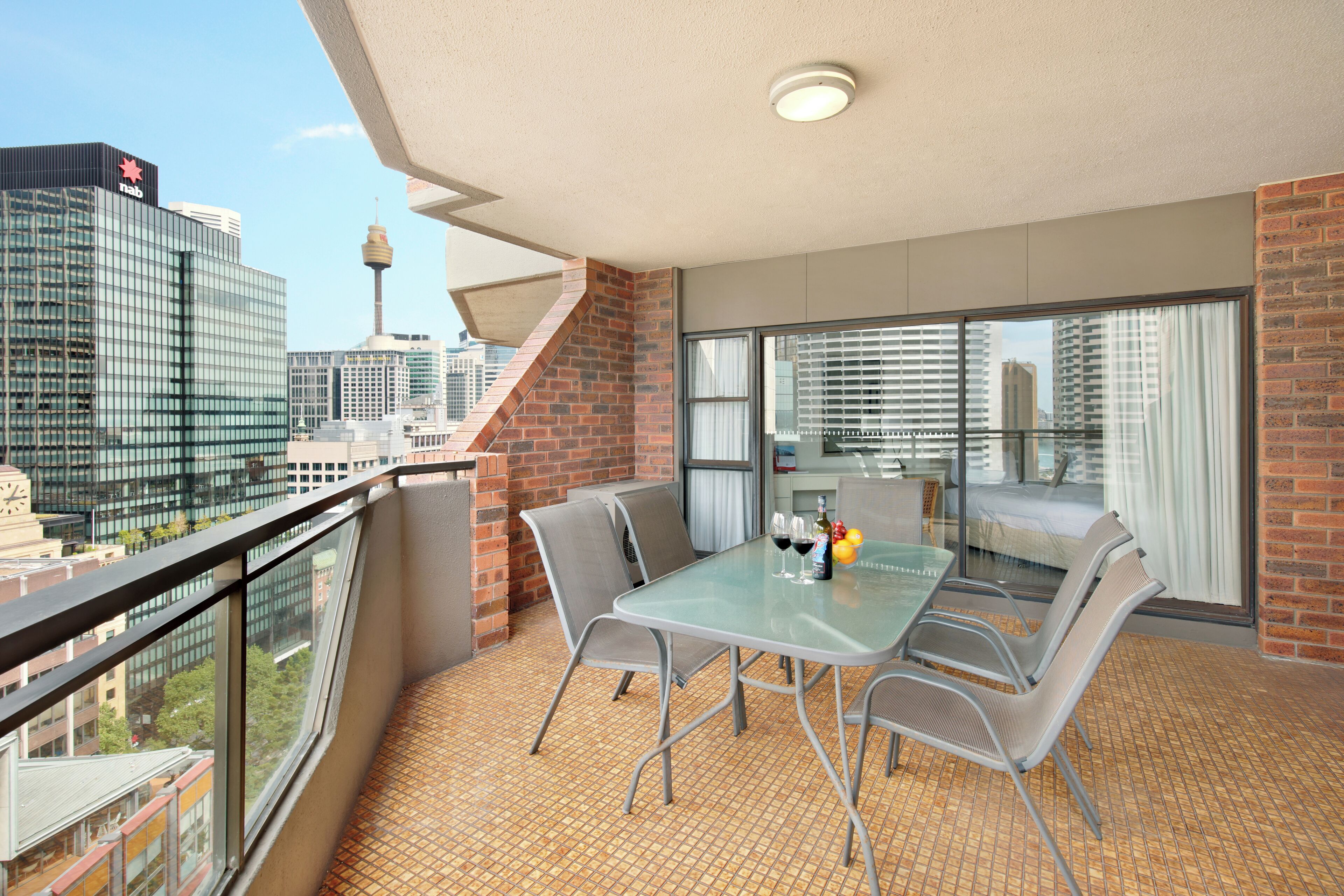 A balcony with a glass table and chairs, a brick wall, and a city view.